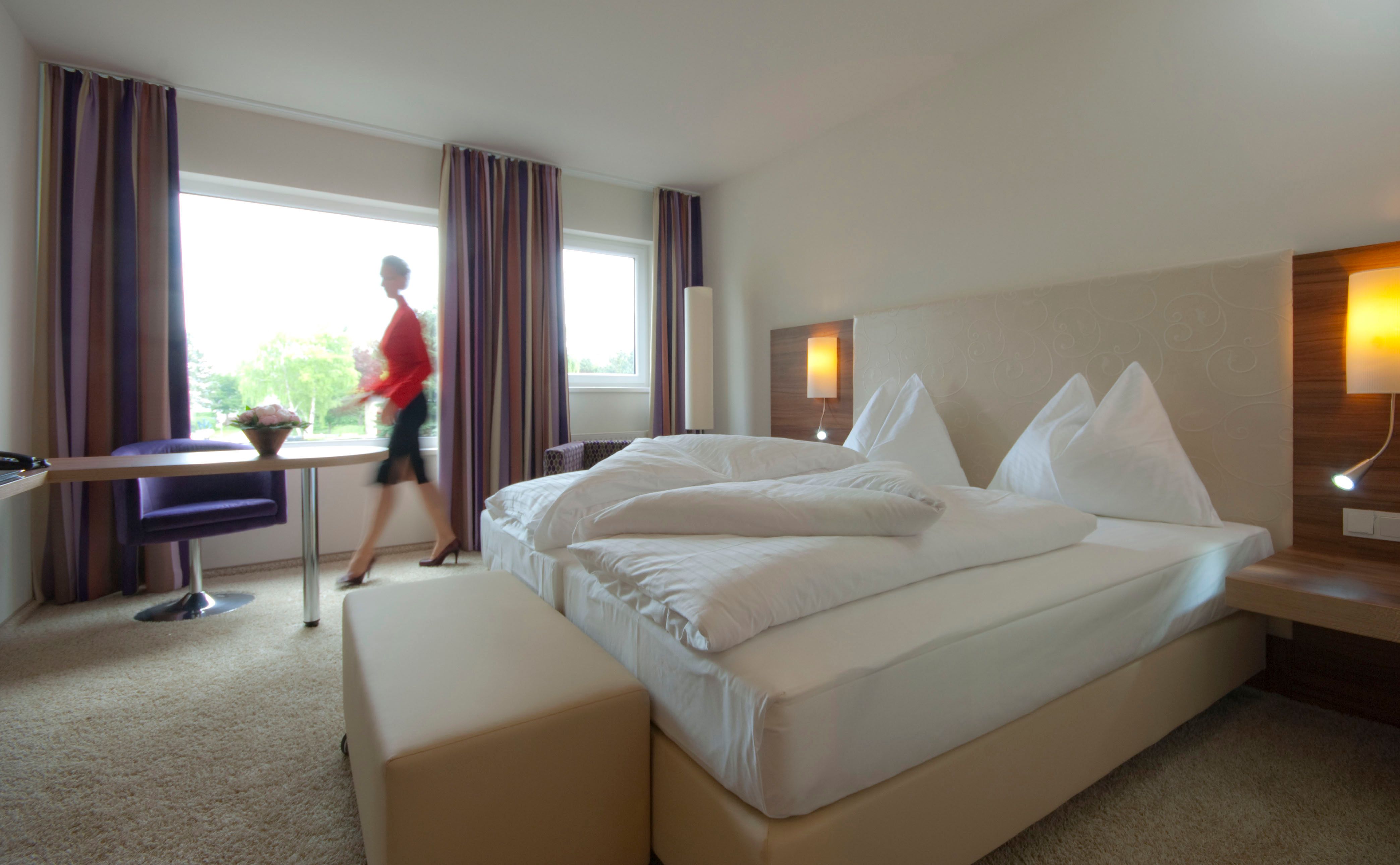 Modern furnished hotel room with double bed, large window and a person walking by.