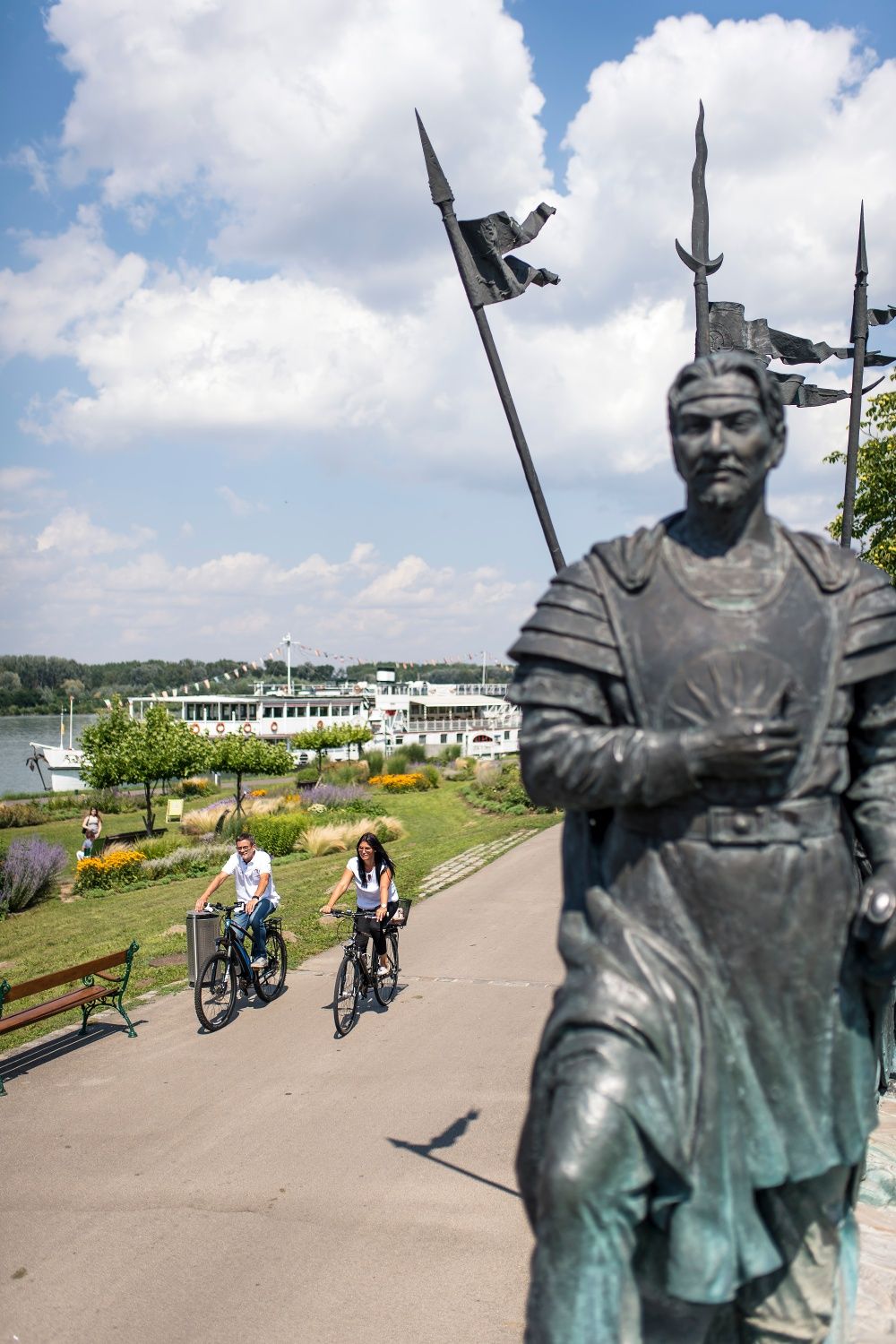 Two cyclists ride on a path along the Danube in Tulln. A statue is in the foreground and a river cruise ship can be seen in the background.