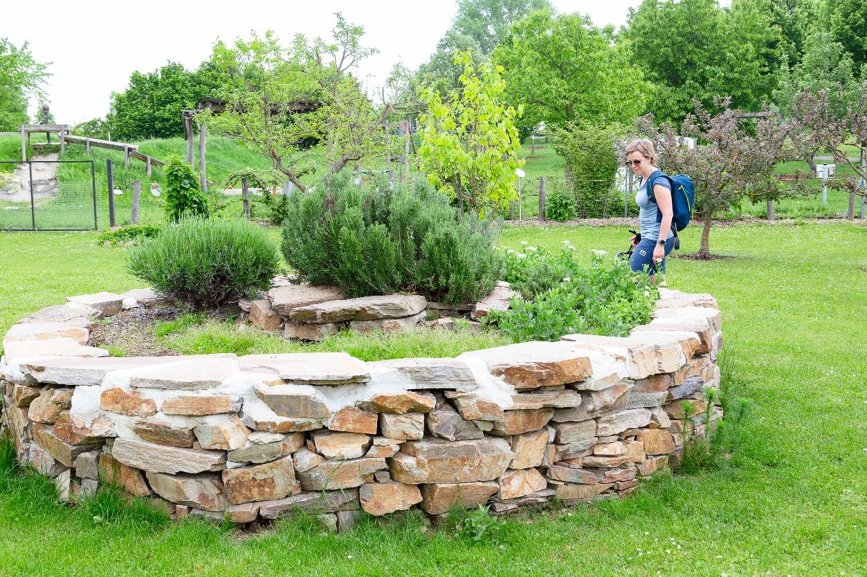 A woman stands next to a herb snail in the Alchemists' Park, surrounded by green vegetation.