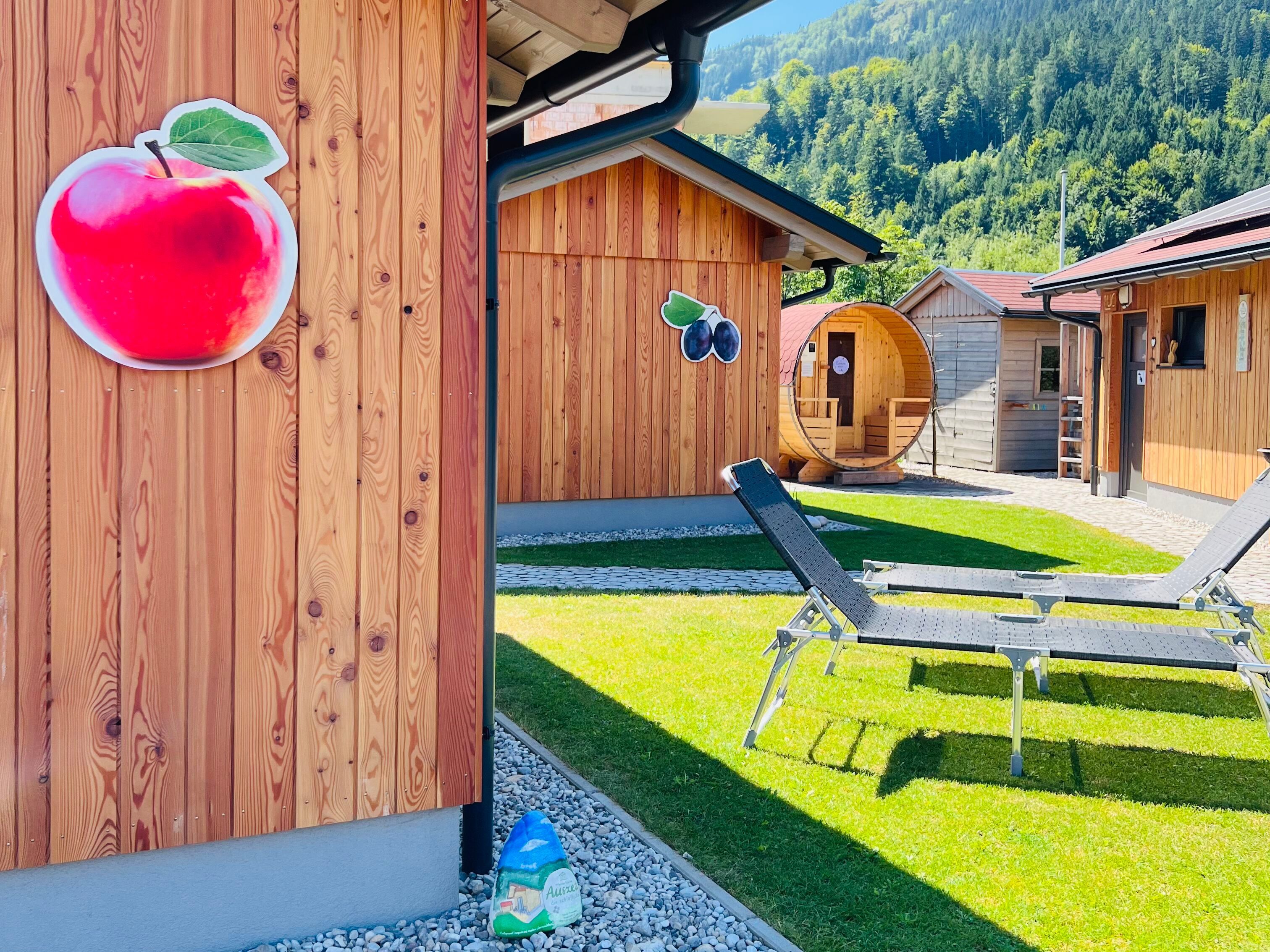 Wooden cabins decorated with fruit and deck chairs on a meadow, surrounded by mountains.