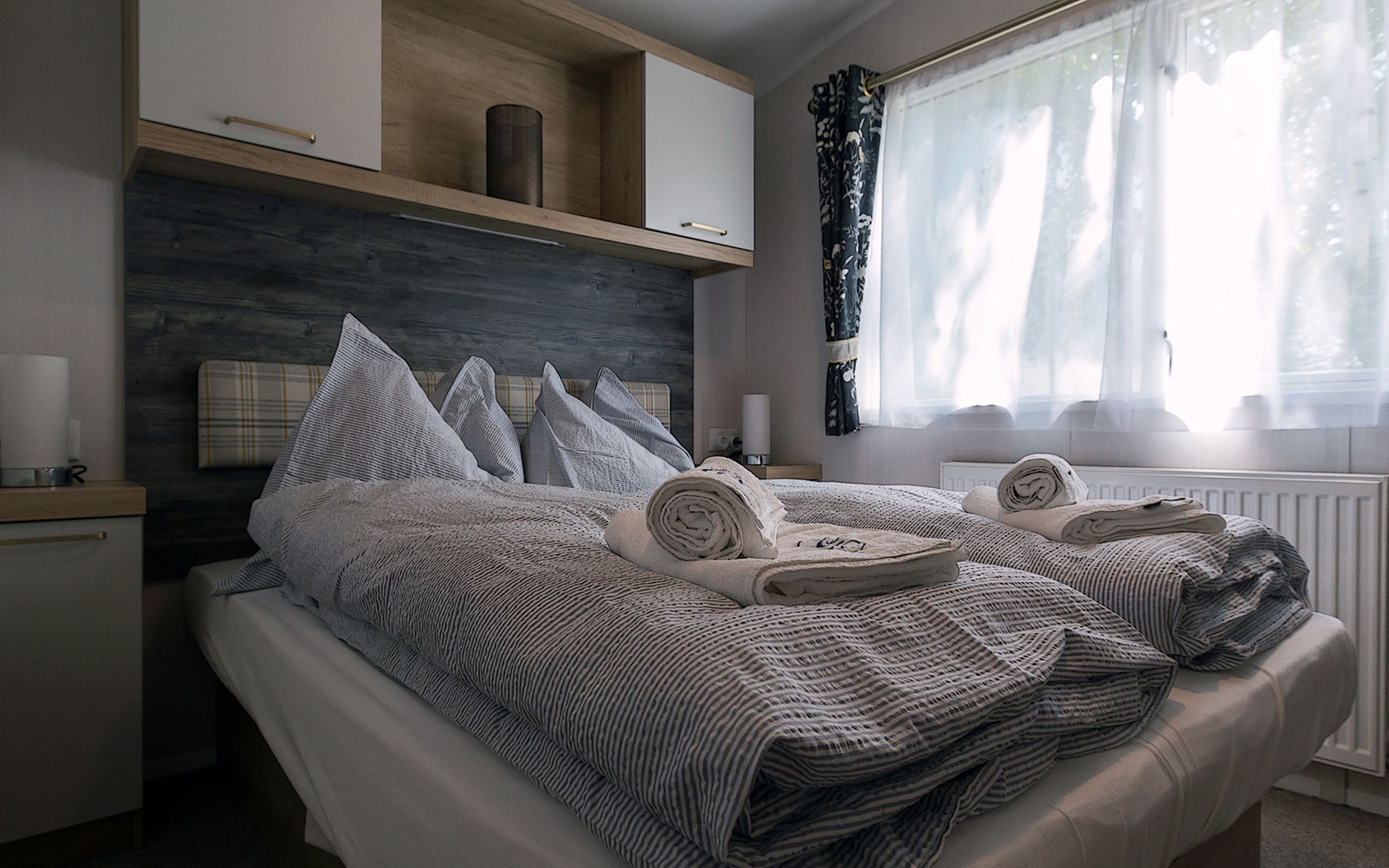 A cozy bedroom with a double bed, grey bed linen and rolled towels. A window with curtains lets in natural light.