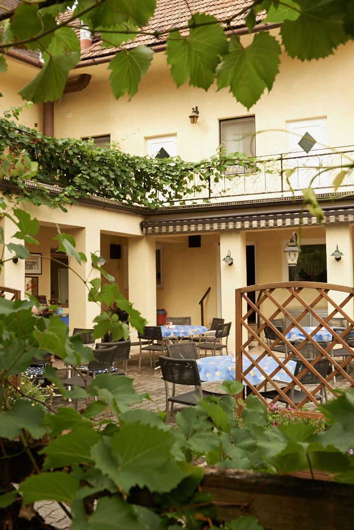 A cozy guest garden with tables and chairs, surrounded by green leaves and a yellow building in the background.