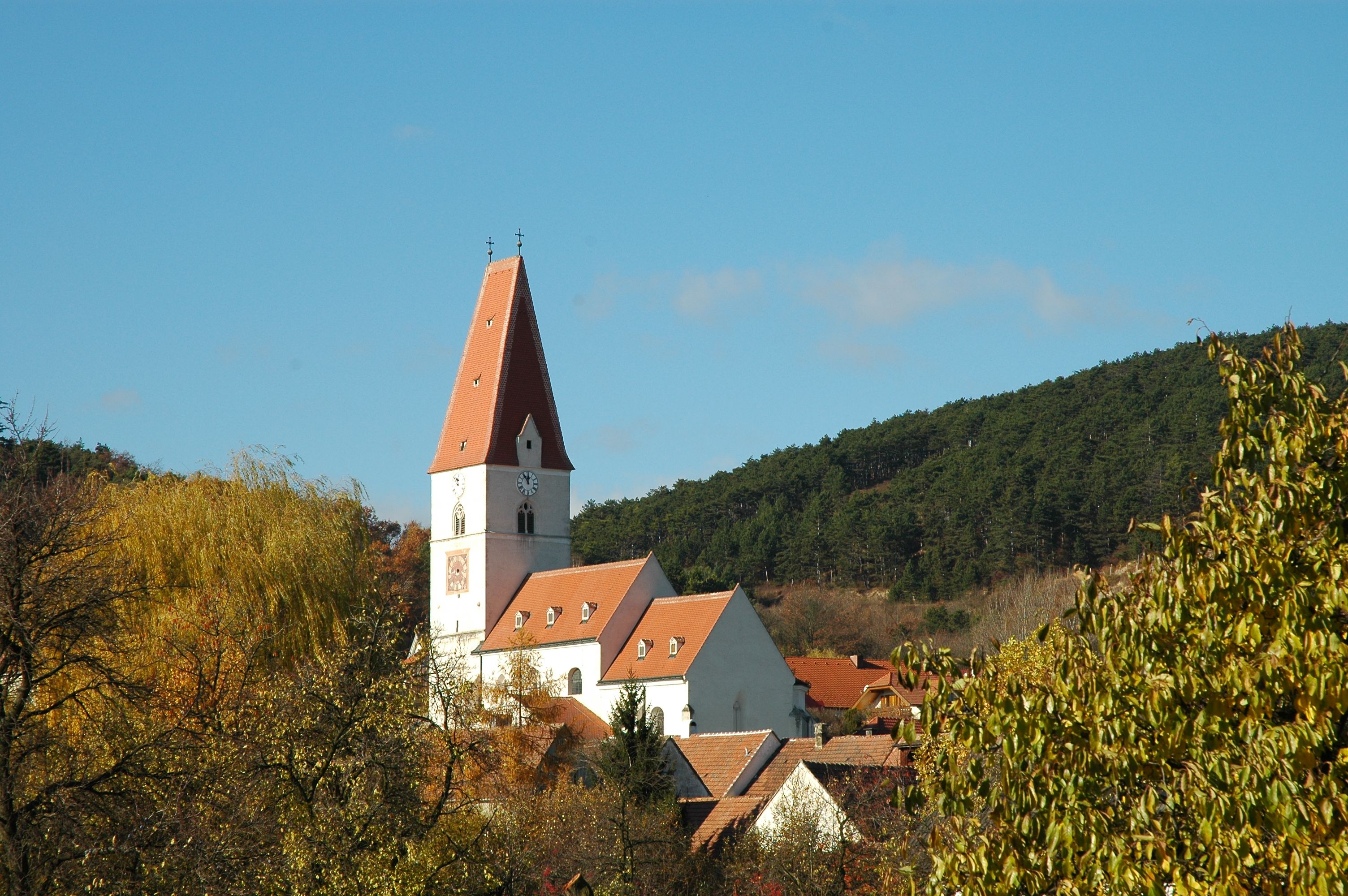 Church in Nußdorf with red roof and clock tower, surrounded by trees and hills.