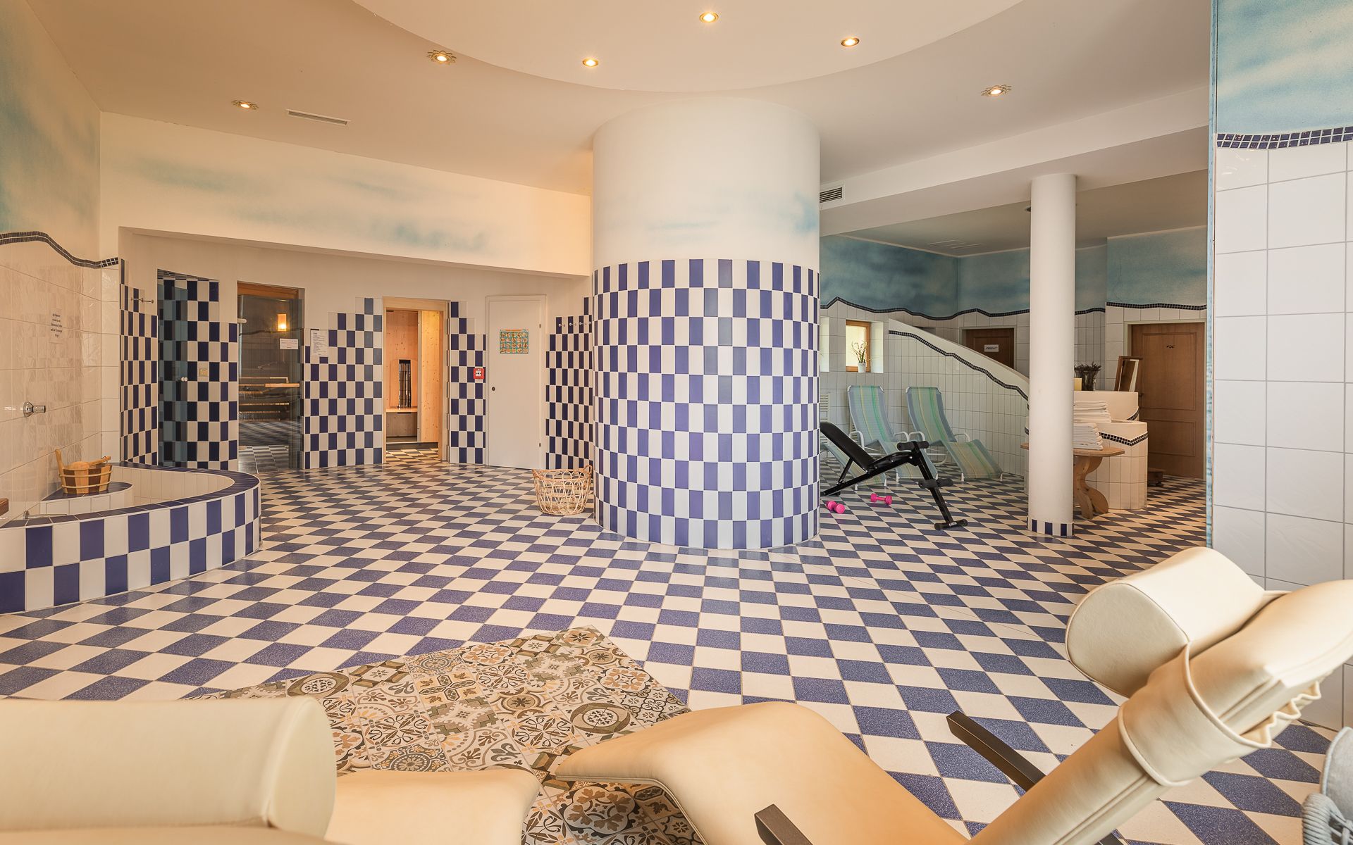 Interior view of a sauna area with blue and white checkered tiles, loungers and fitness equipment.