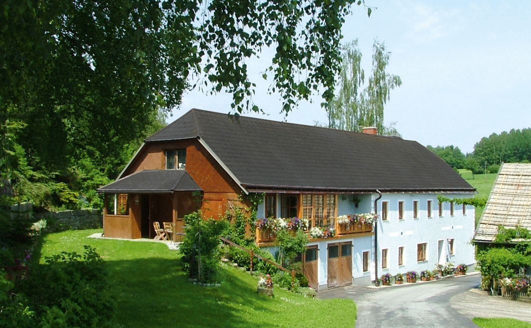 A traditional farmhouse with flowers on the balcony, surrounded by green countryside and trees.