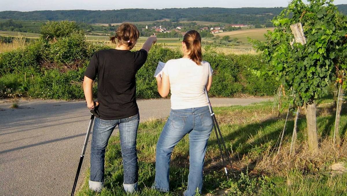 Two women with walking sticks stand on a path and look into the distance, one points in the direction of a village.