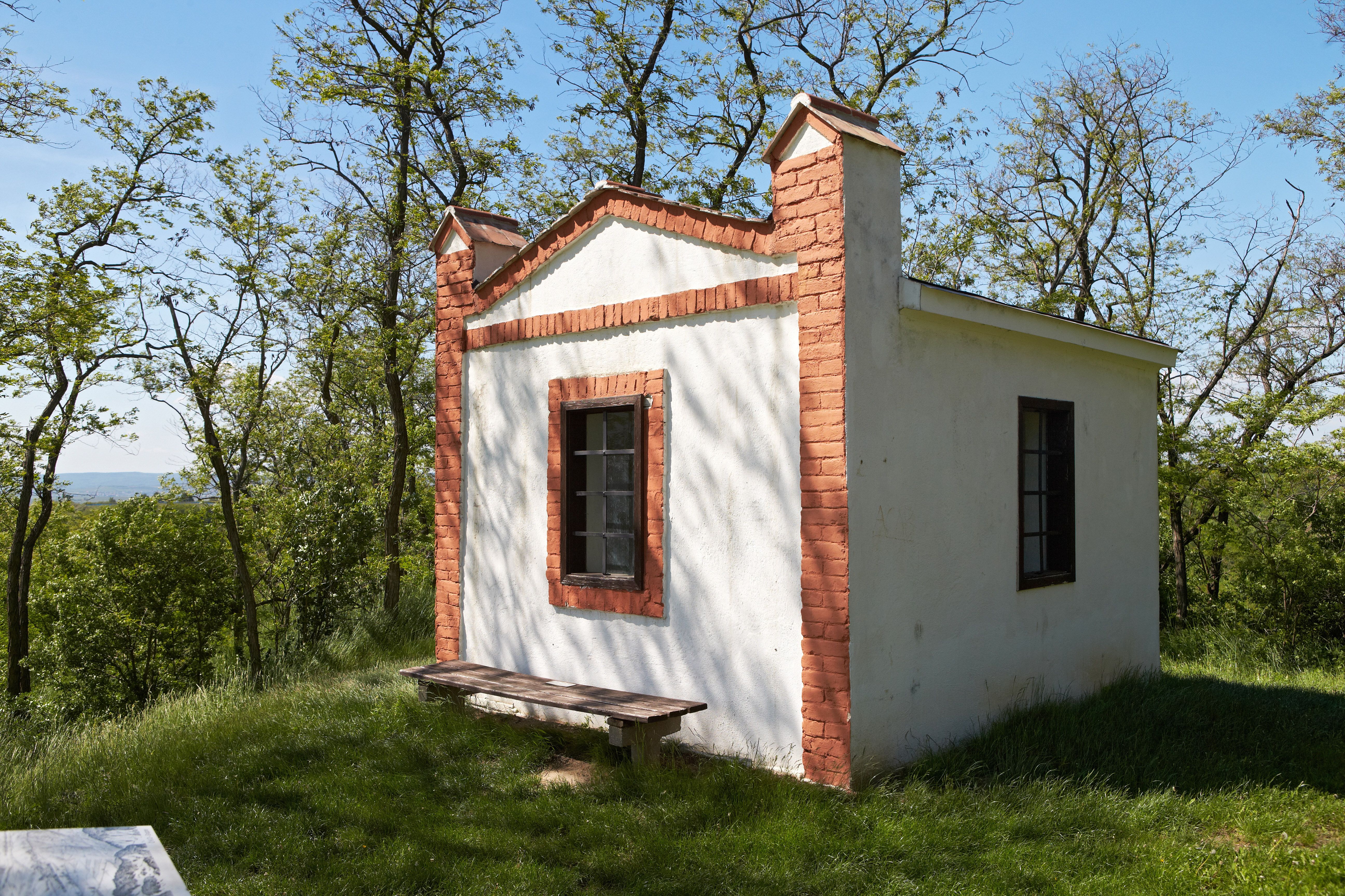 Small white building with red brick accents on a meadow surrounded by trees.