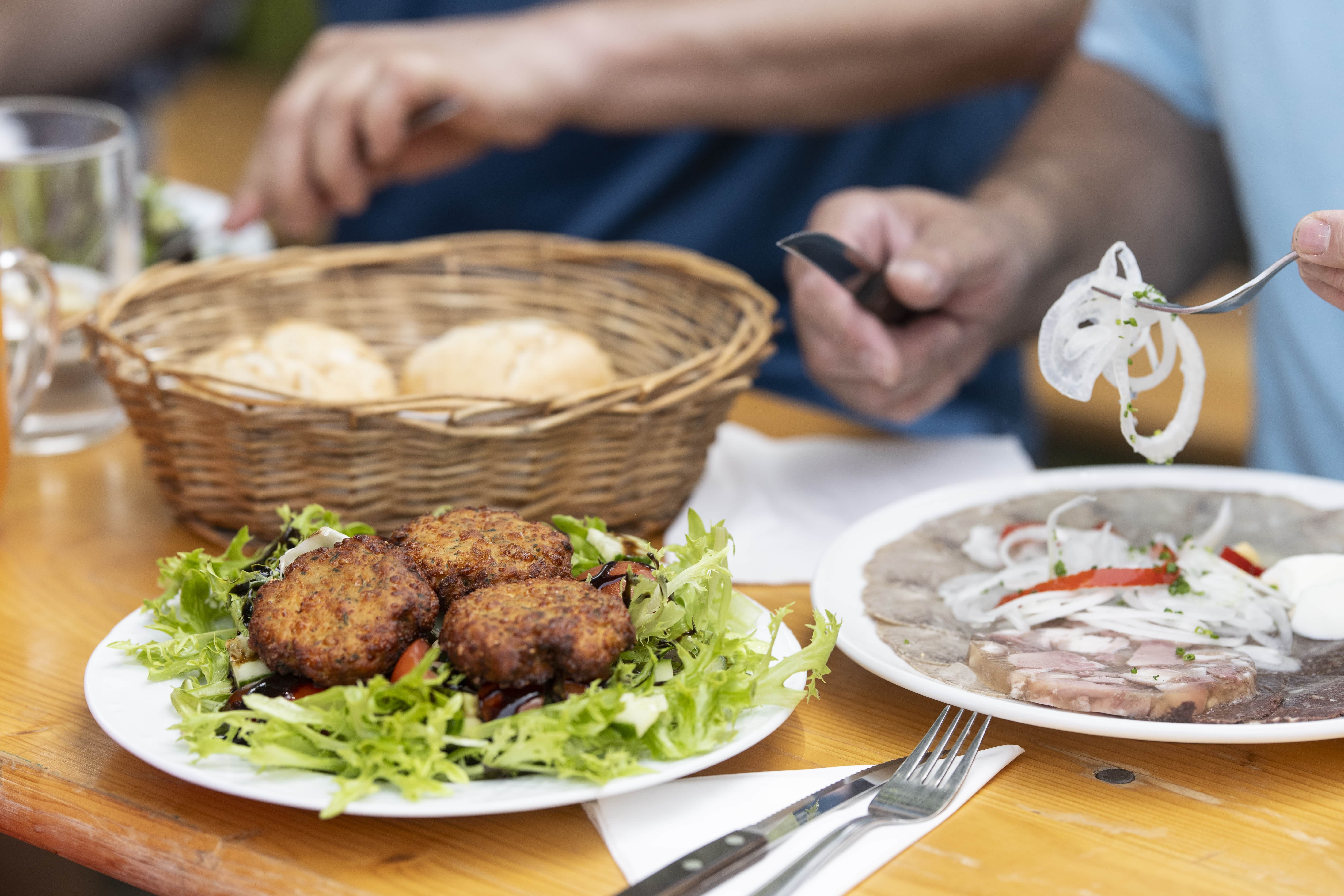 A table with a plate full of meatballs on salad, a basket of pastries and a plate of pressed sausage and onions.
