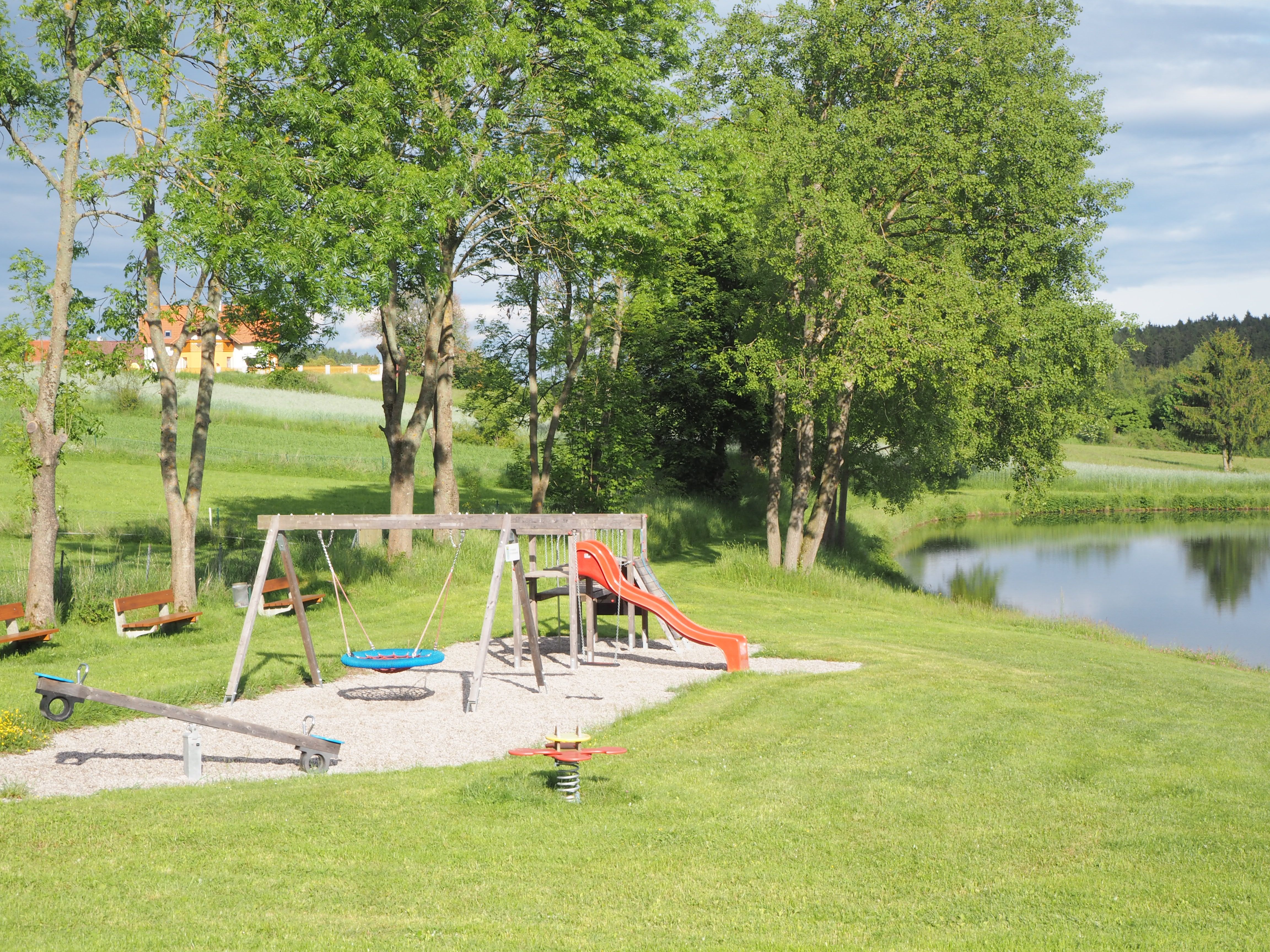 Playground with swing, slide and seesaw next to a pond in a green landscape.