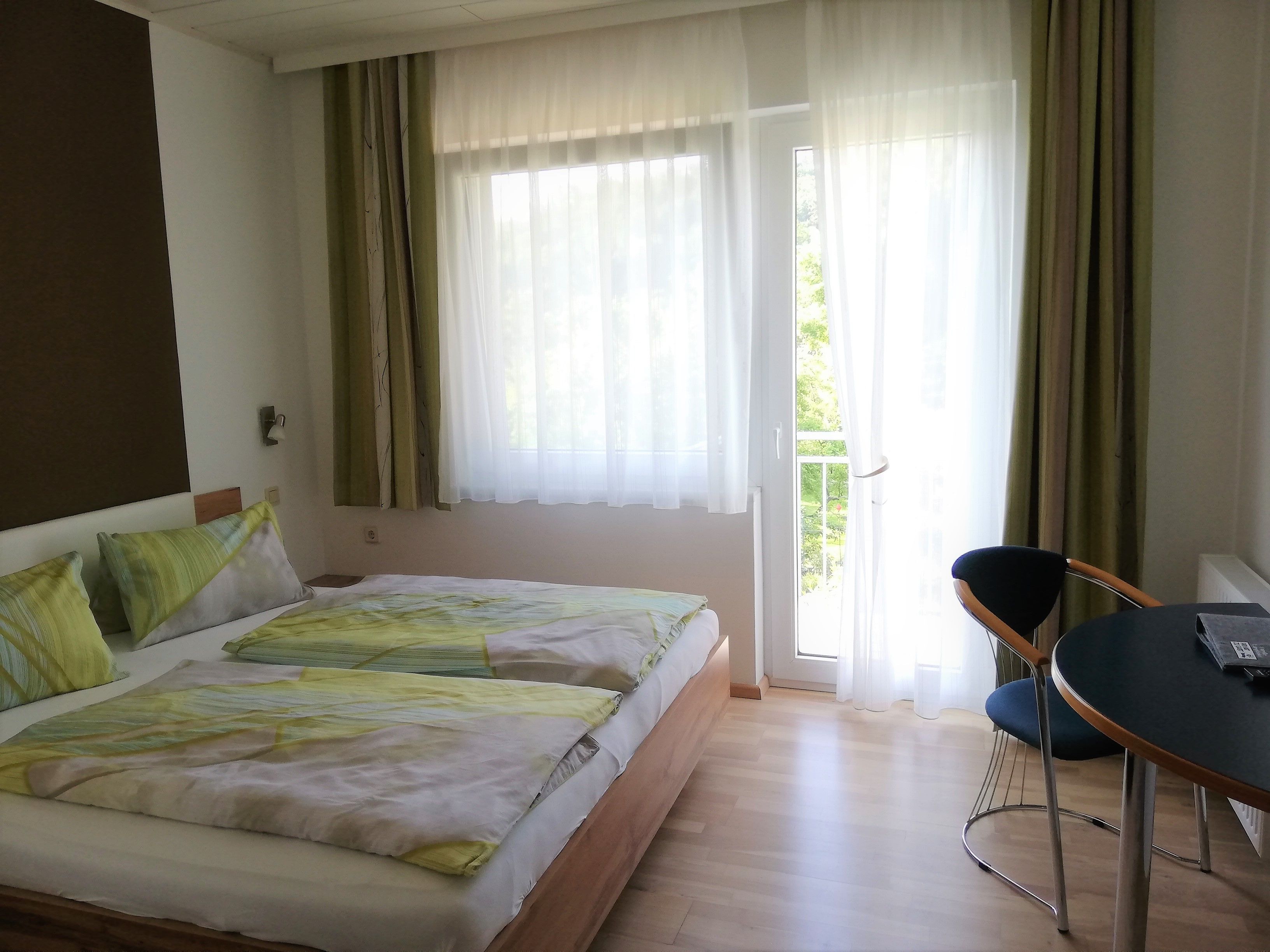 A double room with a bed, a table and a chair. A balcony is visible through a glass door.