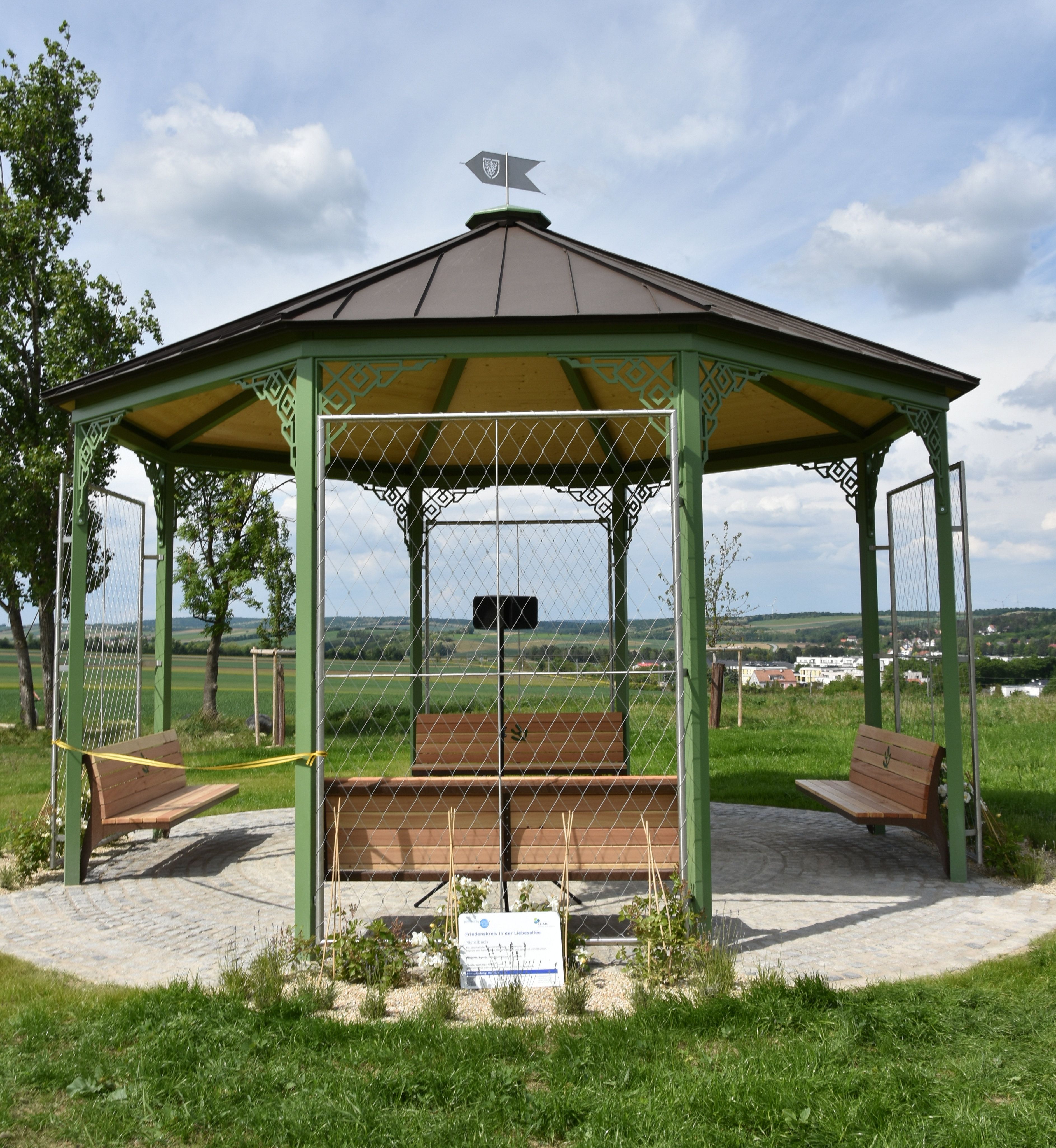A pavilion with a green metal frame and wooden roof stands on a paved square in a rural setting.