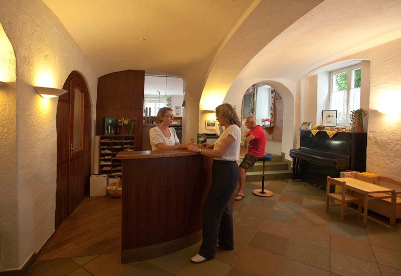 A hotel reception desk with two women in conversation and a man sitting on a stool.