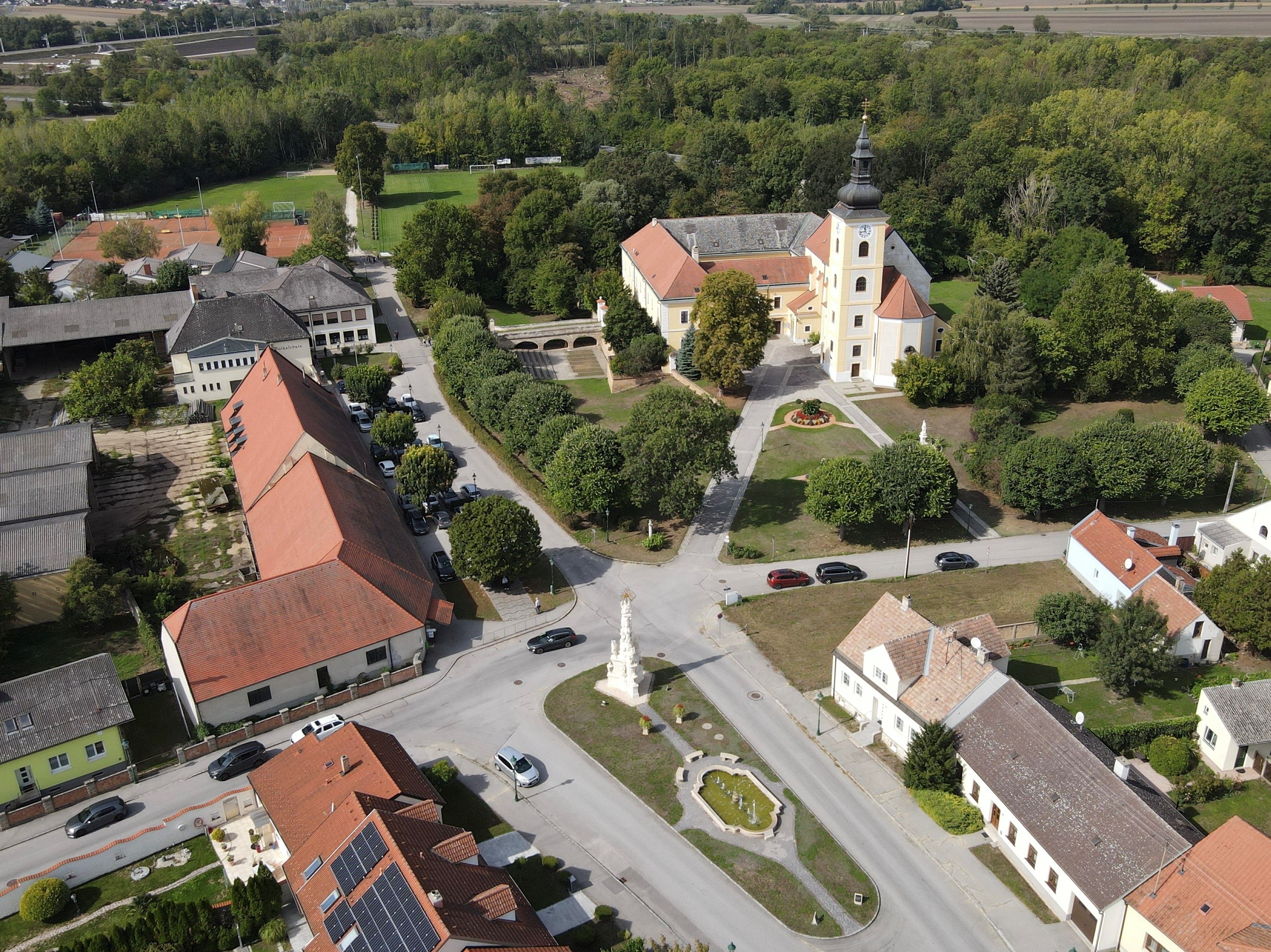 Aerial view of Weikendorf showing the church, village square, fountain, and surrounding houses on the edge of a wooded area.