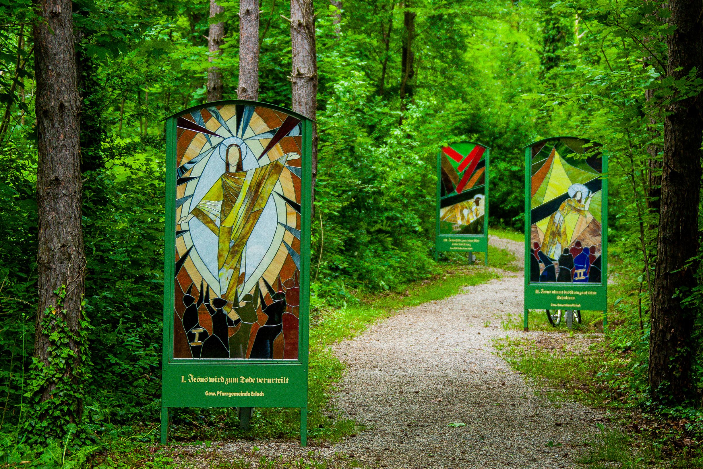 Path through a forest, to the left and right of the path are large glass panels depicting the Stations of the Cross.