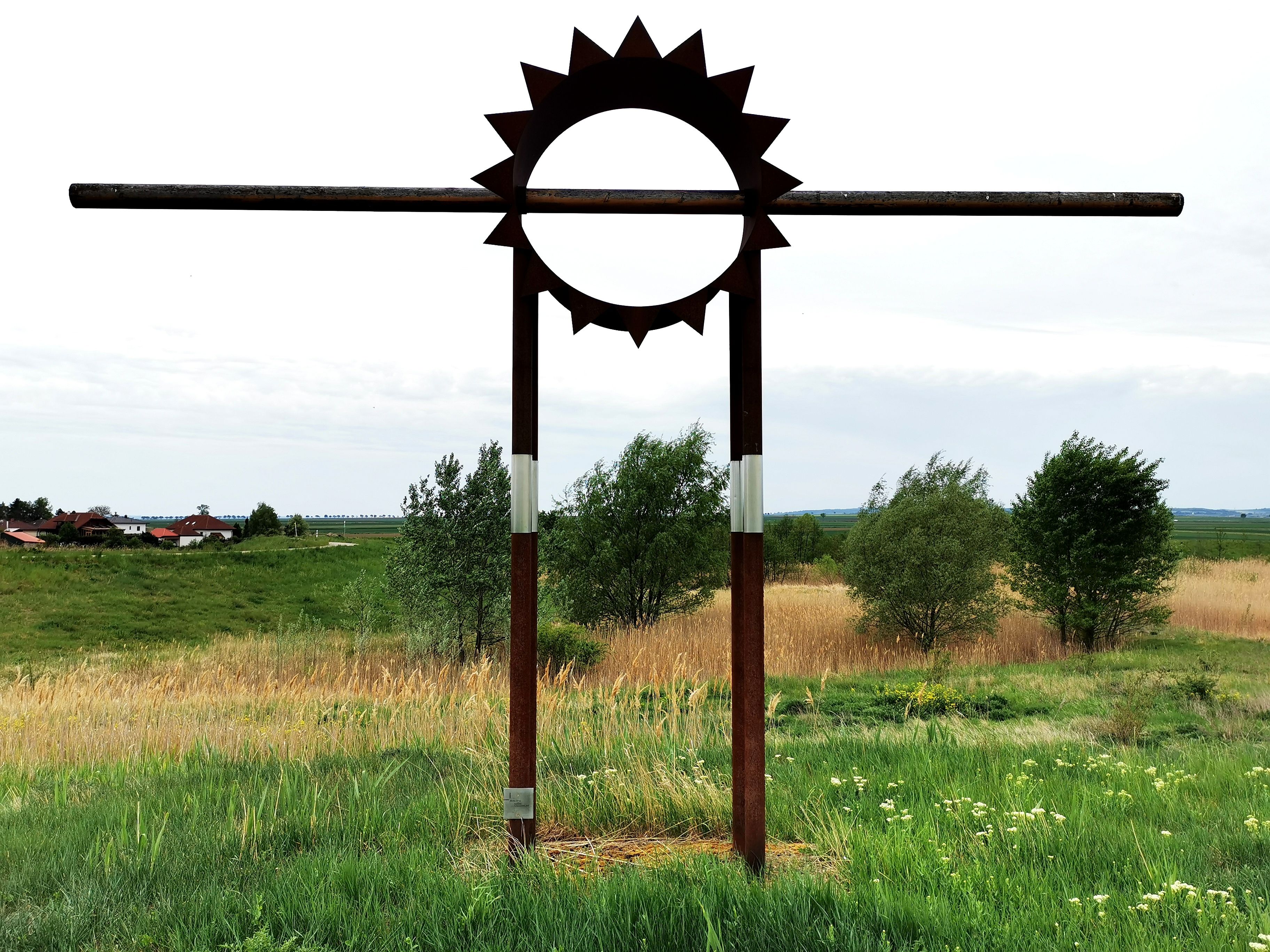 Metal sculpture in the shape of a sun on a meadow with trees in the background.