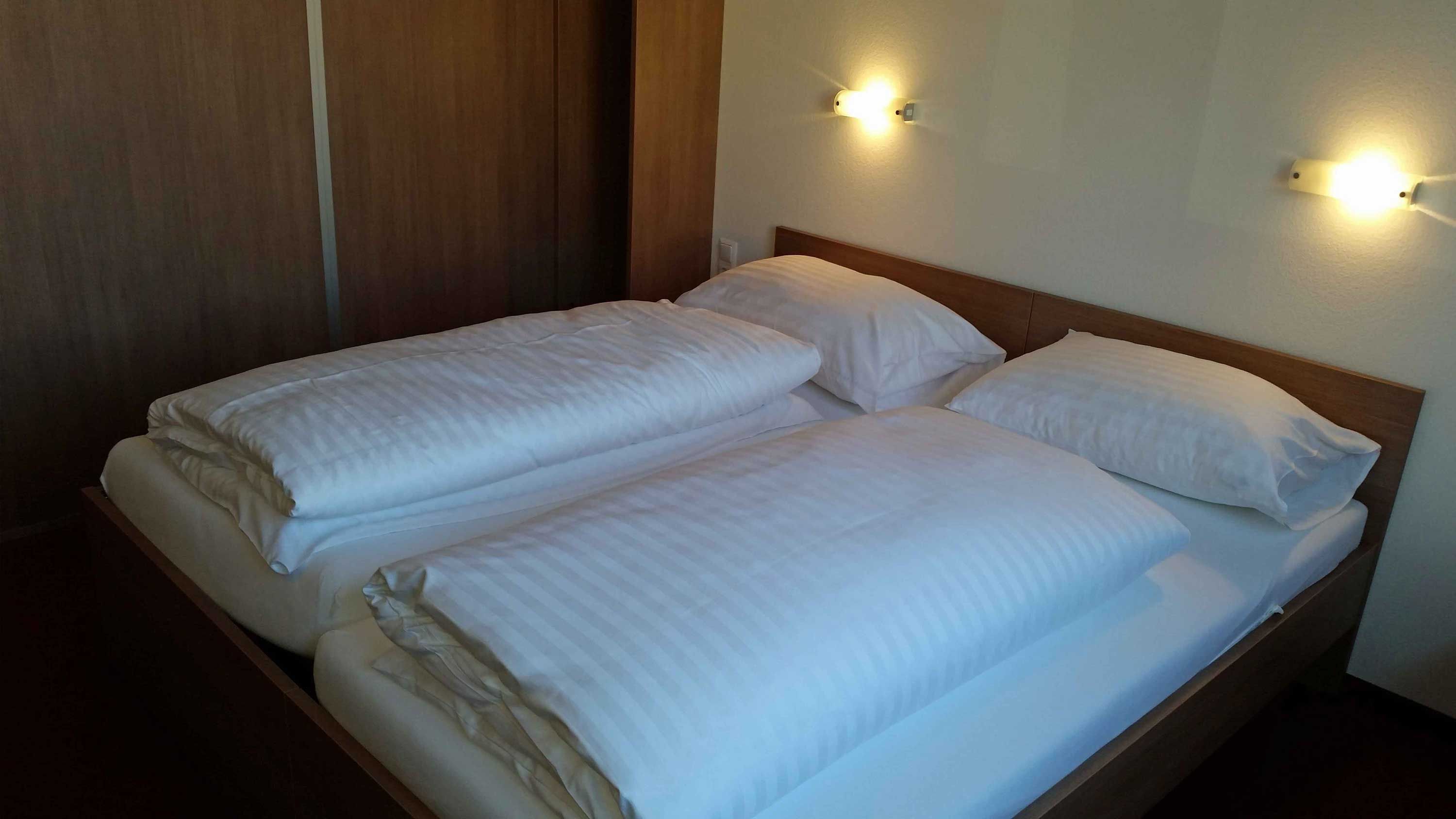A double bed in a motel room with white comforters and pillows, illuminated by two wall lamps.