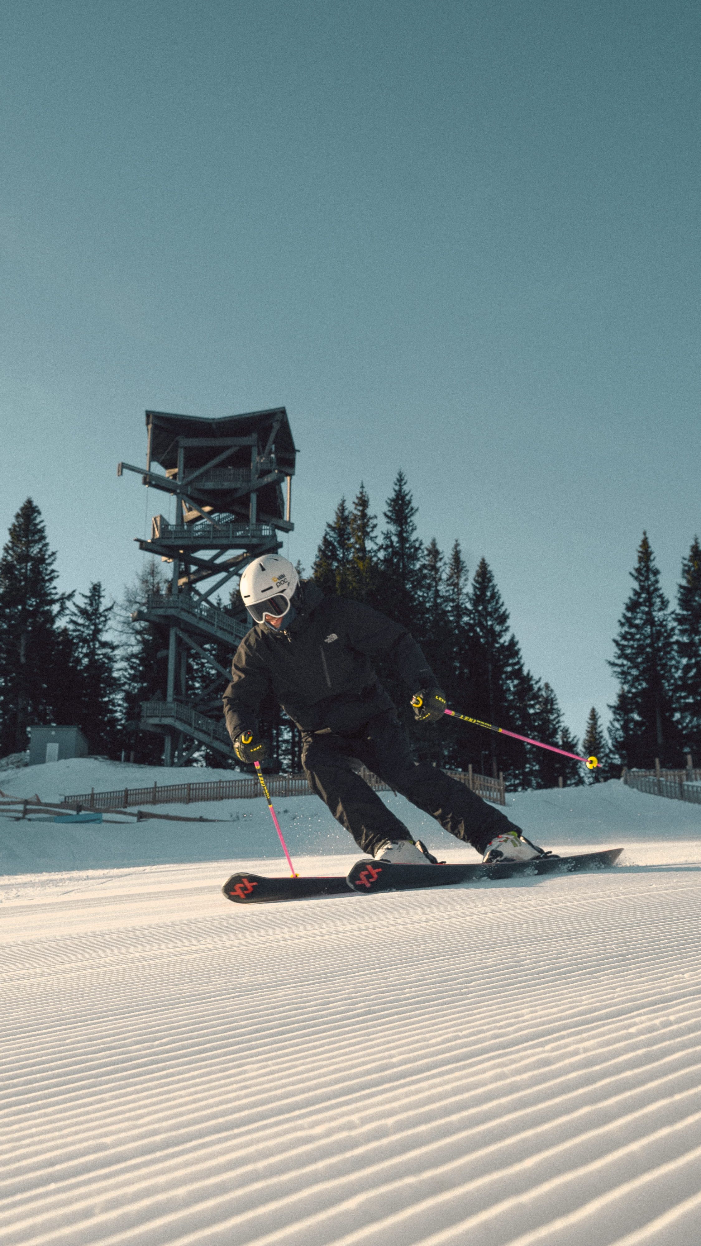 Skier in black clothing skiing on a groomed slope on the Semmering Hirschenkogel, with a lookout tower and trees in the background.