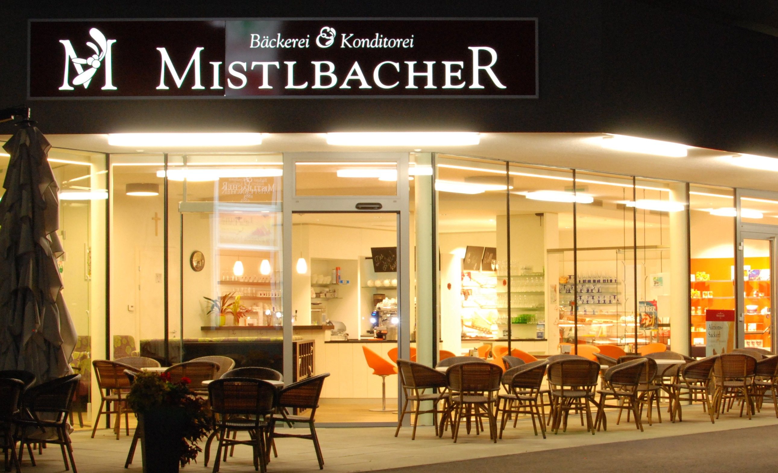 Exterior view of a café called Mistelbacher, illuminated and with outdoor seating.
