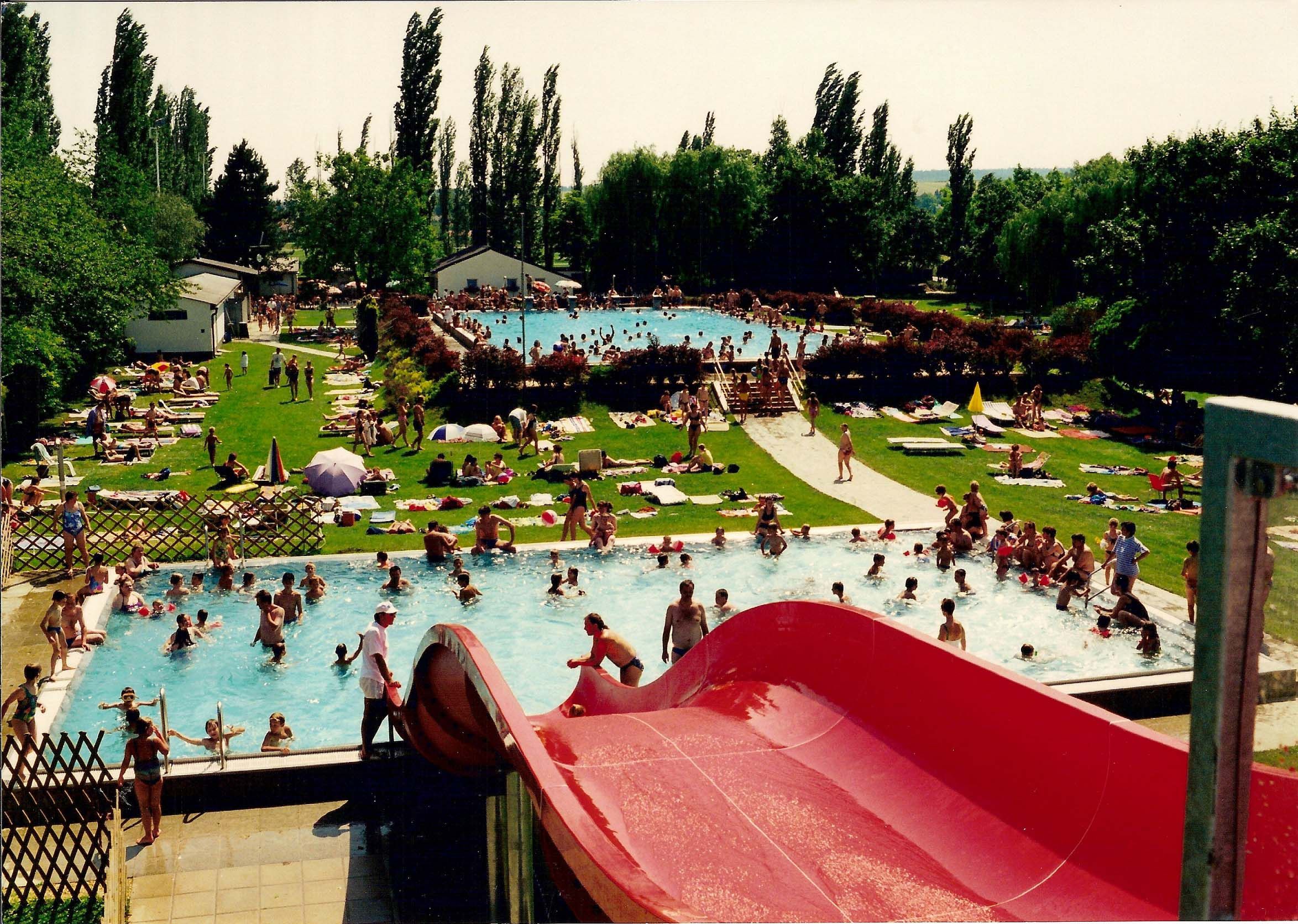 An outdoor pool with several pools, a red water slide and lots of people swimming in the water or lying on the grass.