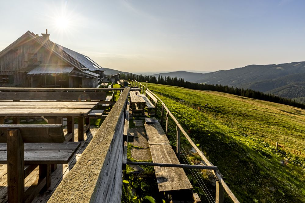 A mountain hut with wooden benches and tables, surrounded by green meadows and mountains in the sunlight.