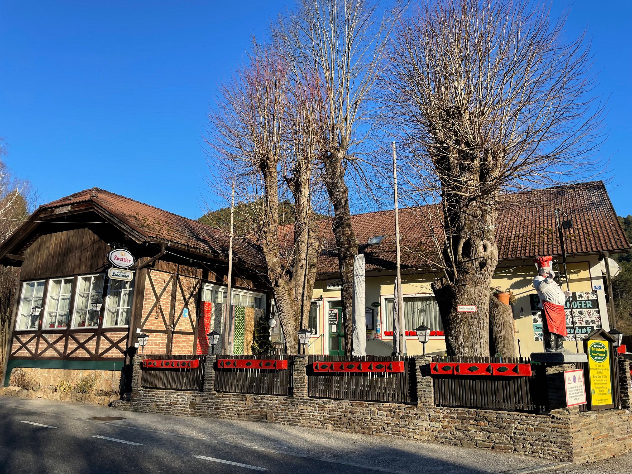 Half-timbered building with red roof tiles, surrounded by bare trees and a fence. A figure dressed as a cook stands in front of the building.