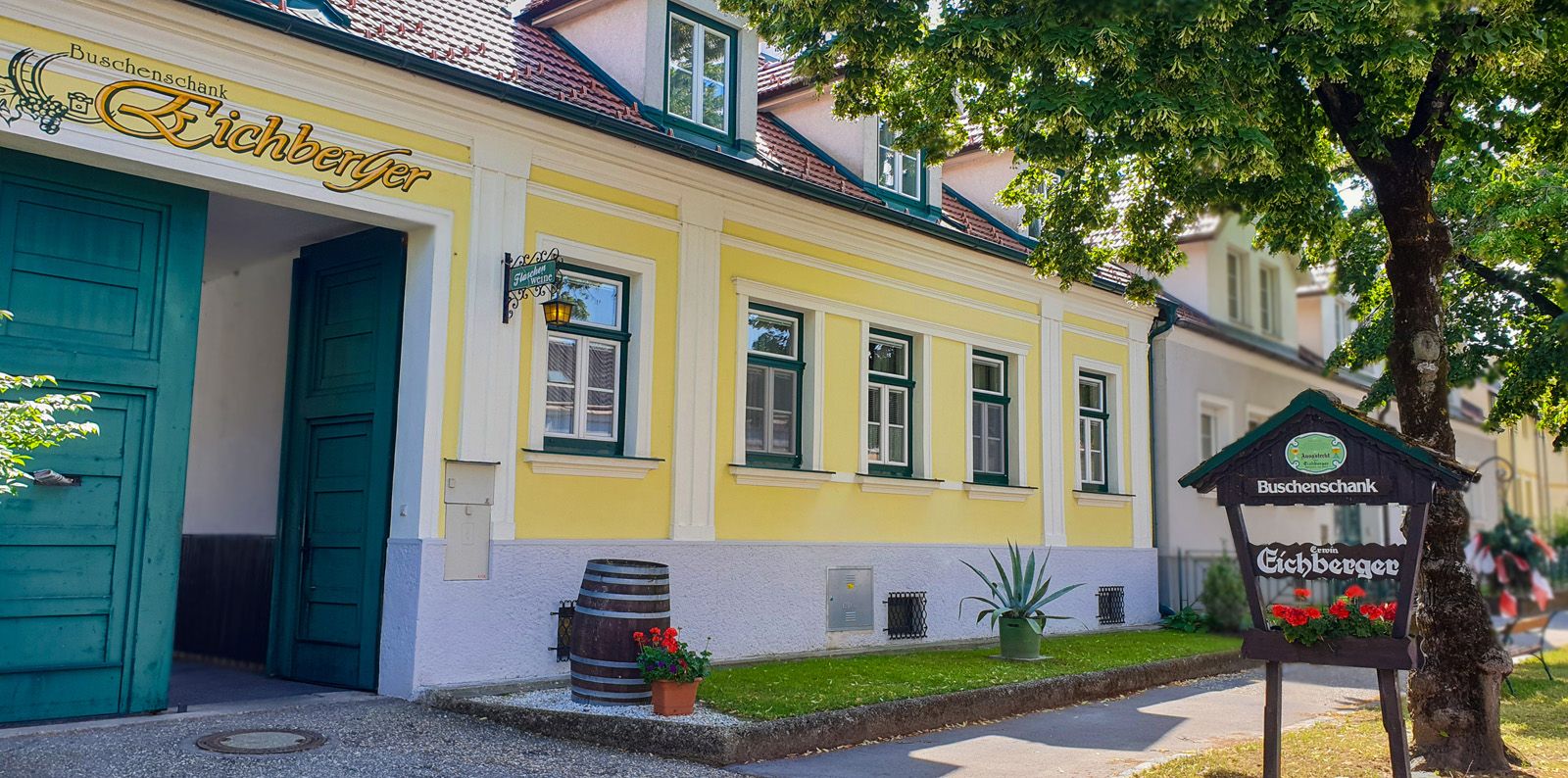 Yellow building with green doors and windows, sign 'Buschenschank Eichberger', tree in the foreground.