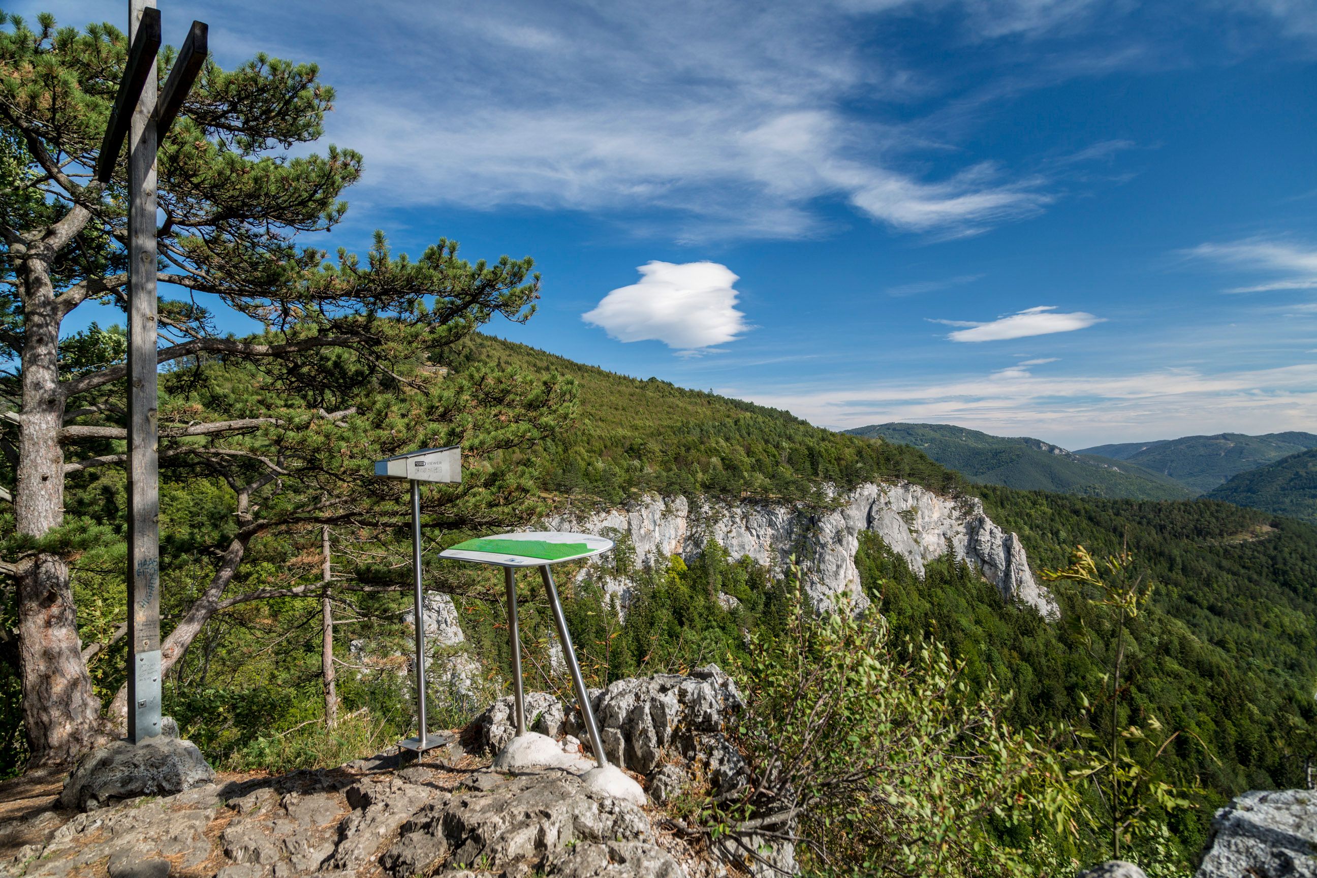 Viewpoint with information board and cross on a rock, surrounded by forest and mountains under a blue sky.