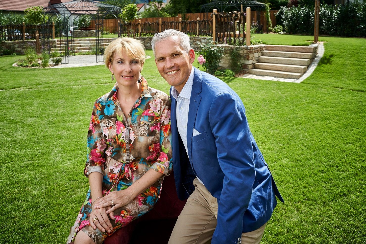 The Wildeis family are sitting in a meadow, smiling at the camera. The man is wearing a blue jacket, the woman a colorful dress.