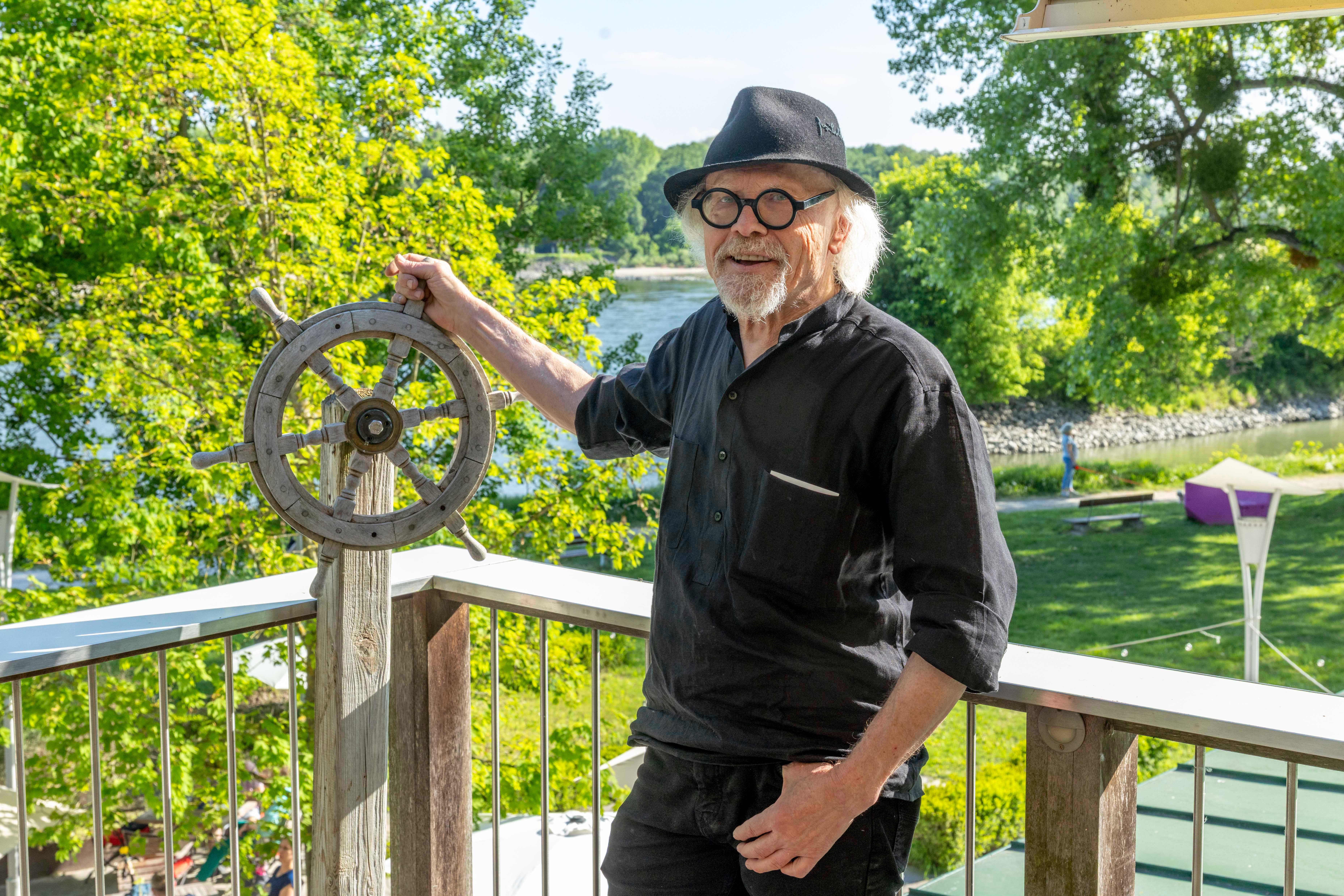 A man with a hat and glasses is standing on a balcony holding a steering wheel. Trees and a river (the Danube) can be seen in the background.