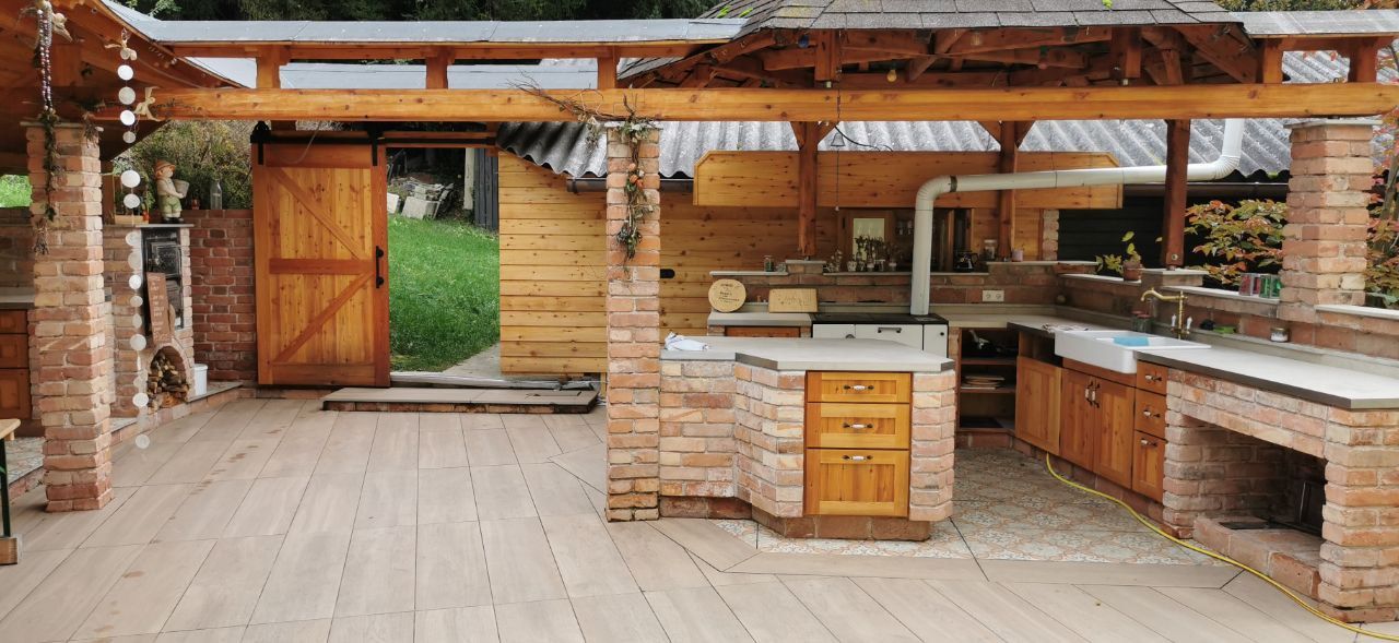 Open outdoor kitchen made of bricks with wooden furniture and wooden roof and with access to a green meadow.
