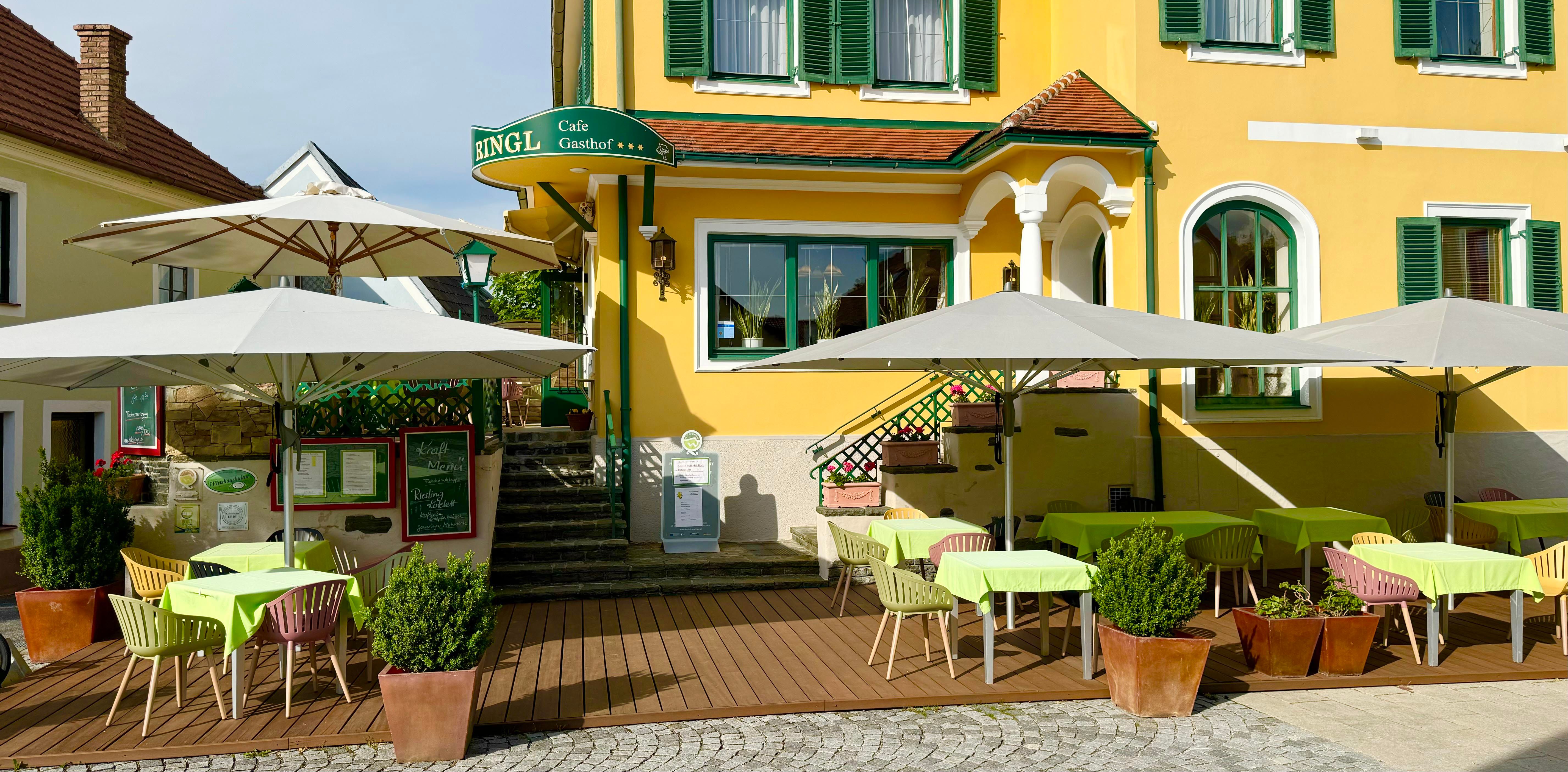 A yellow building with green shutters and a garden with tables and parasols.