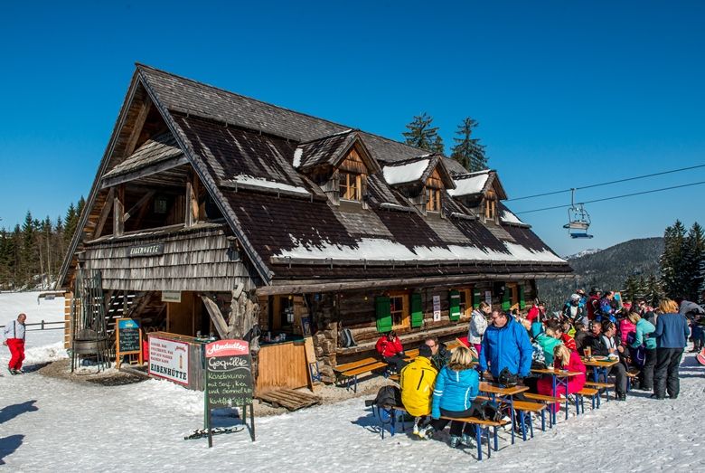 A mountain hut in the snow with people sitting outside.
