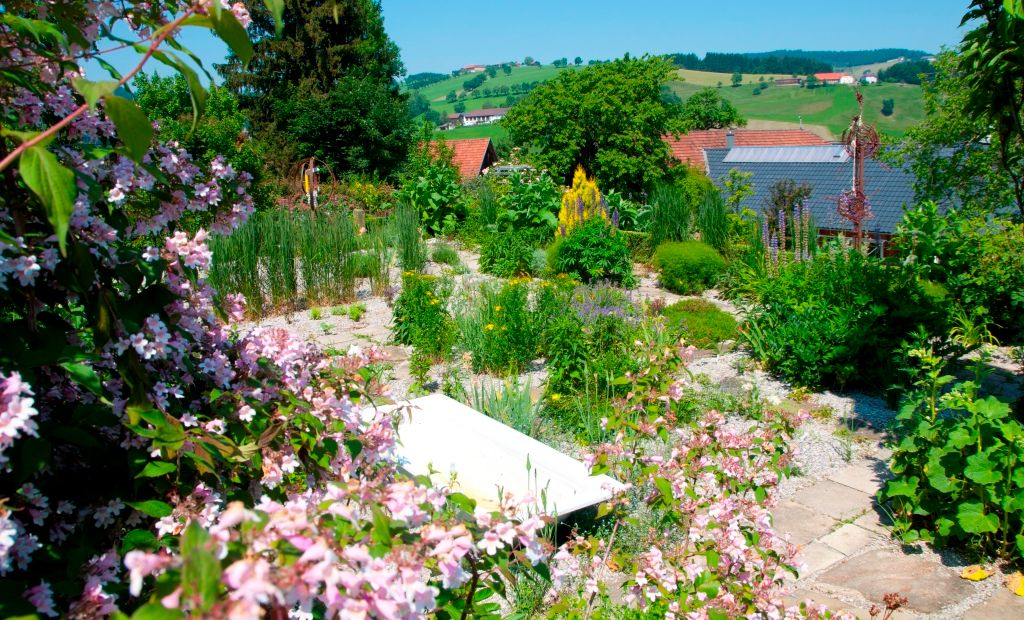 A blooming garden with colorful flowers and green plants, hills and a house roof in the background.