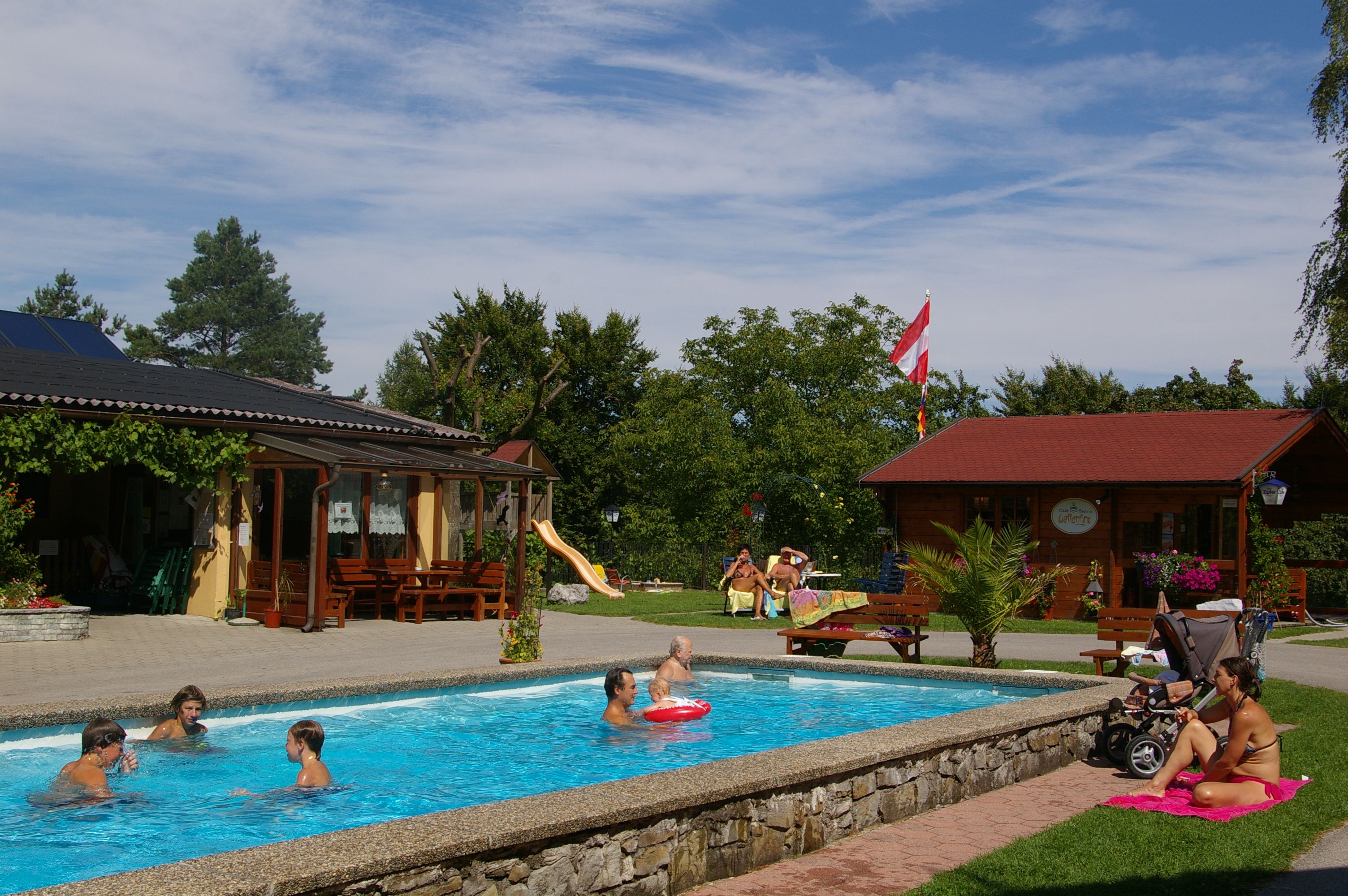 People relax by a small pool in a garden with wooden houses and a slide.