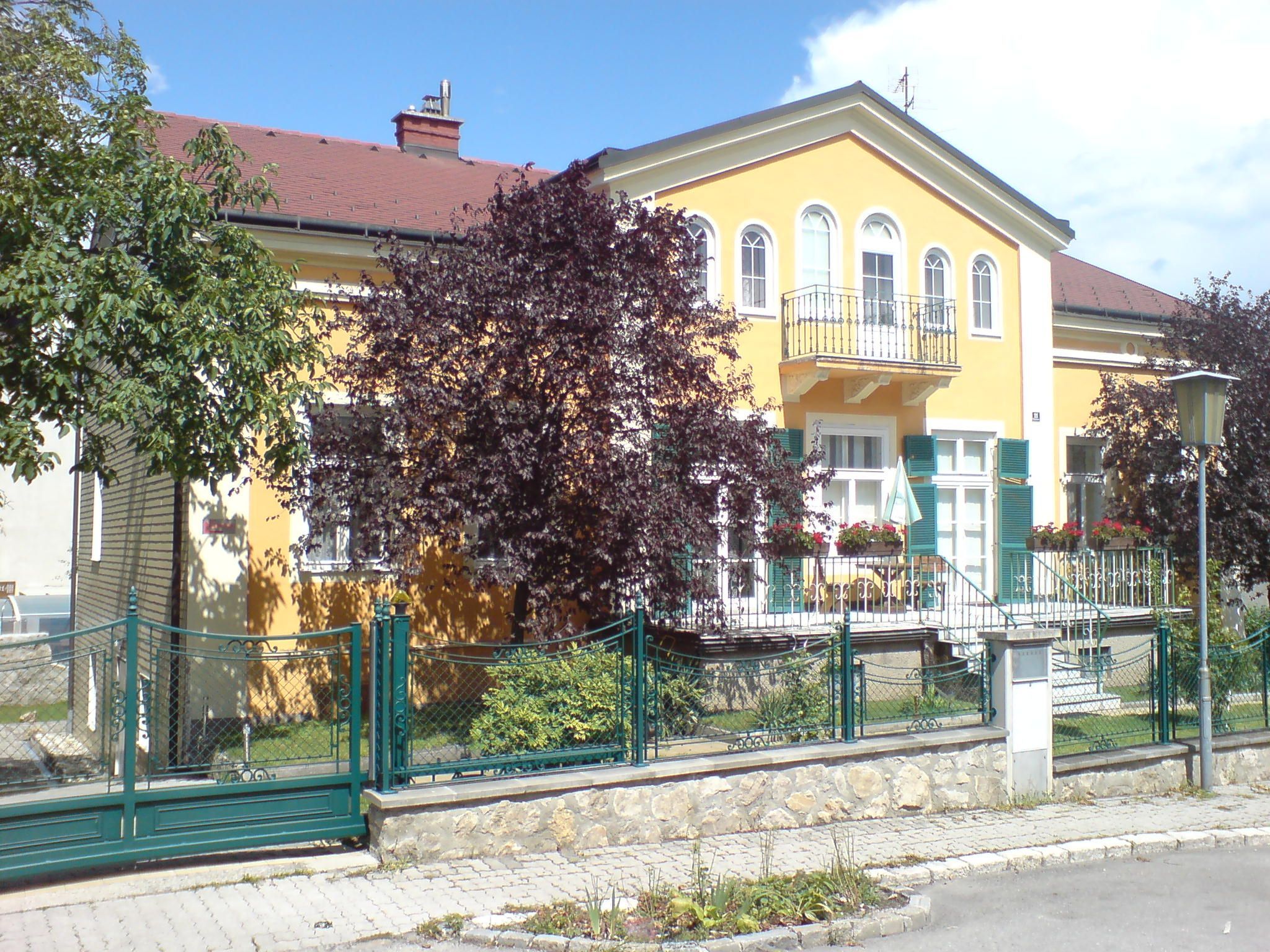 Yellow house with balcony and green fence, surrounded by trees.