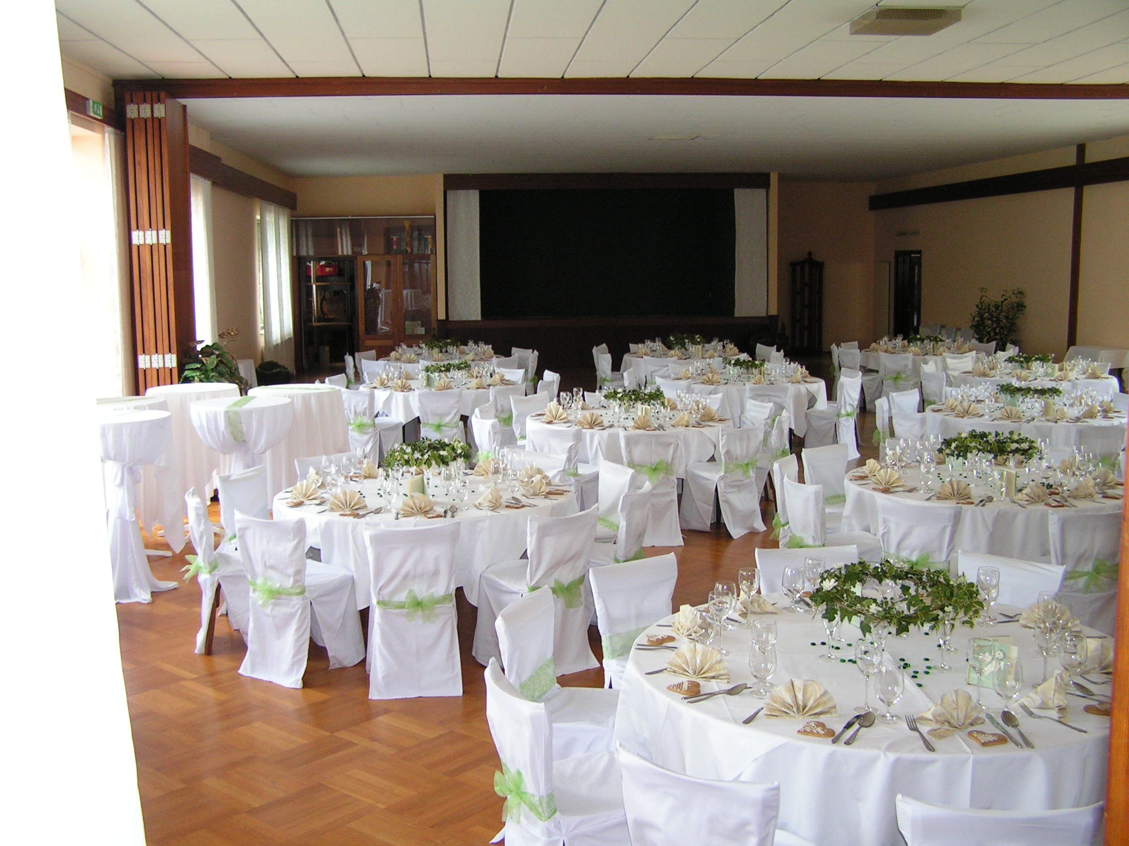 Elegant wedding hall with round tables, white tablecloths and chair covers, decorated with green plants and folded napkins.