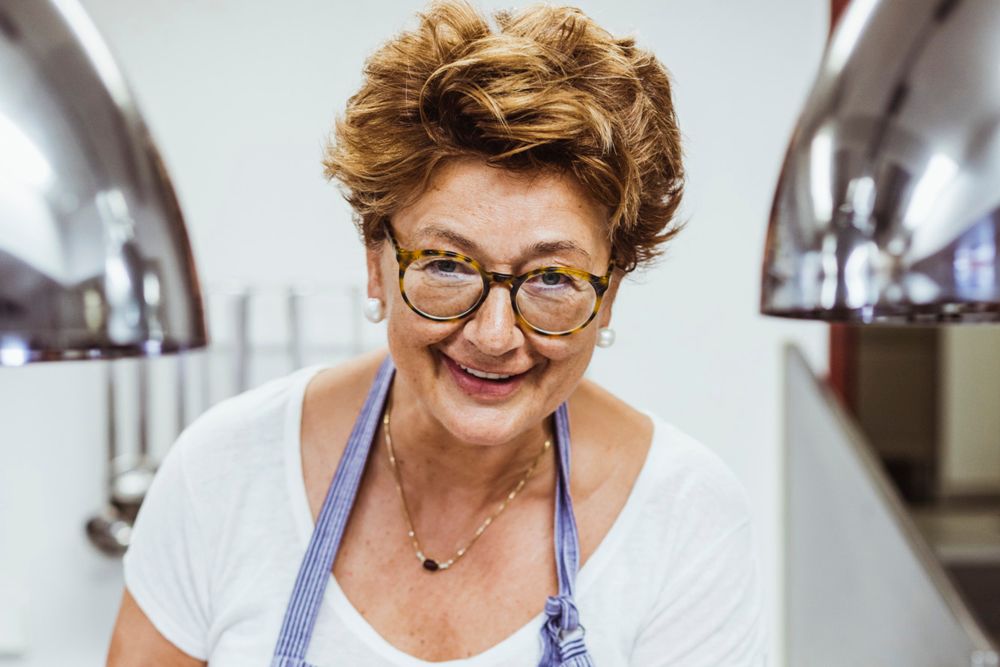 A smiling woman with glasses and an apron in a kitchen.