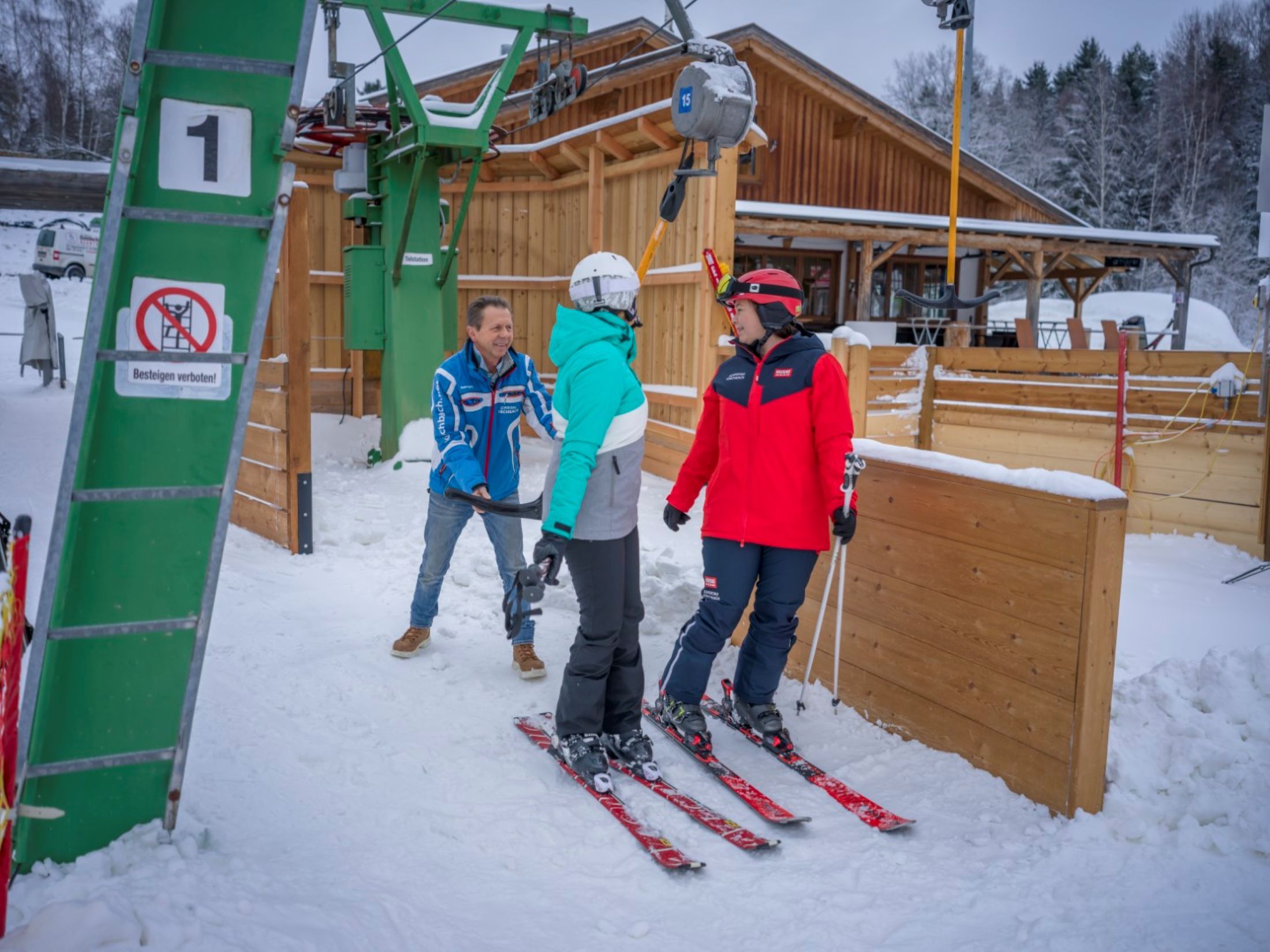 Two female skiers stand at a ski lift in a snowy setting while a man helps them.