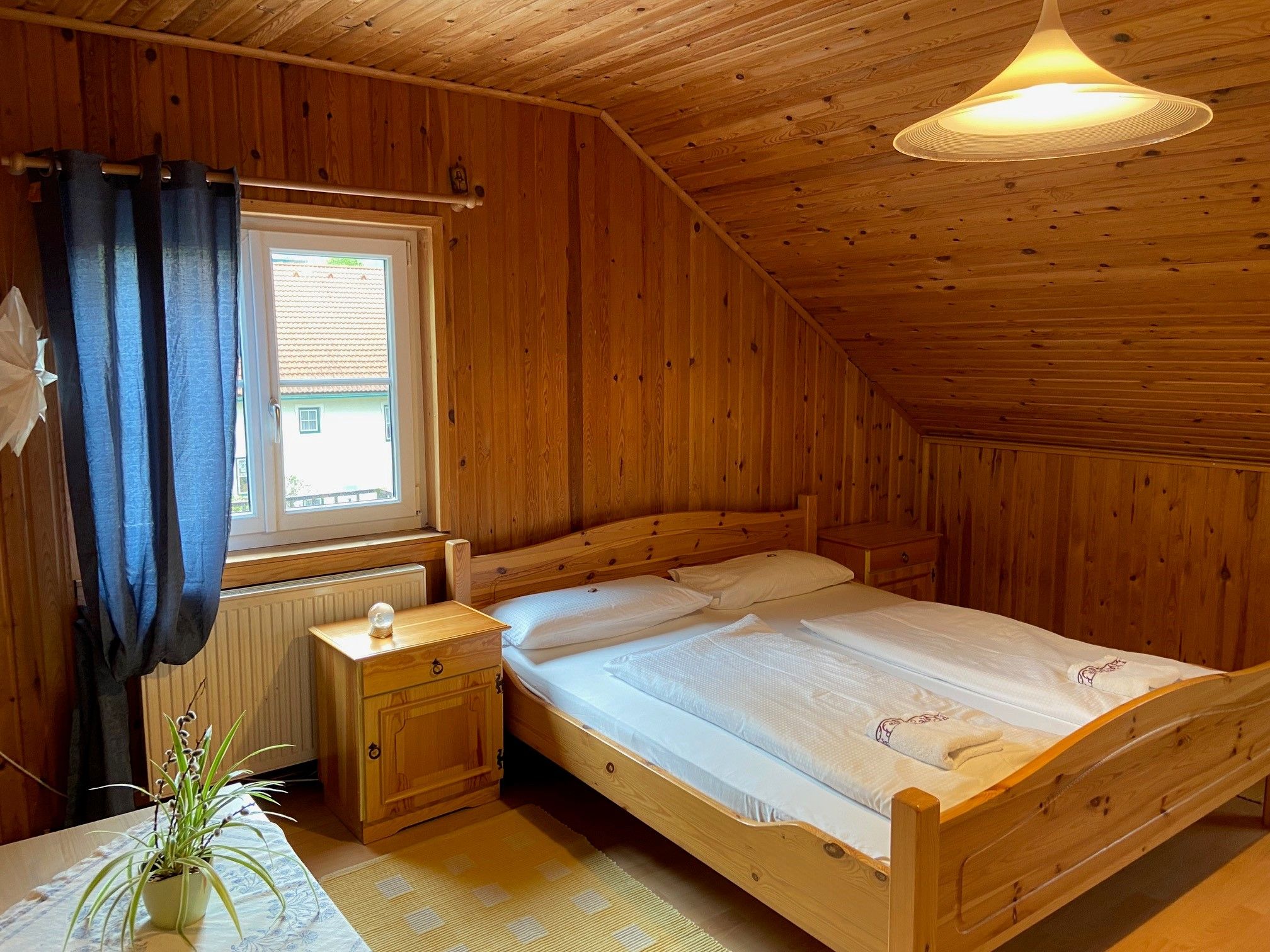 Cozy double room with wood paneling, double bed, window with blue curtains and a plant on the table.