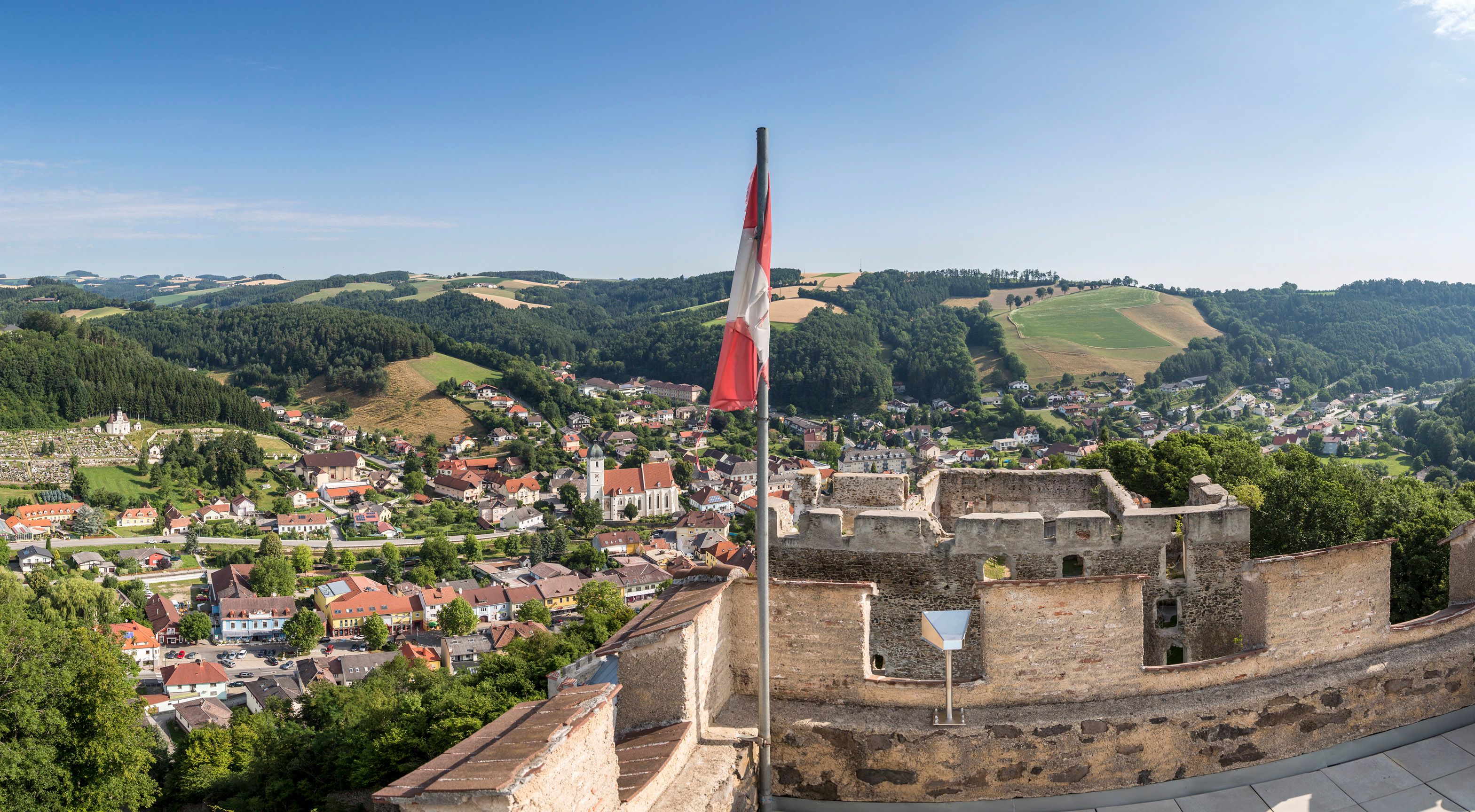 Panoramic view from a ruined castle to a town with hills in the background and an Austrian flag in the foreground.