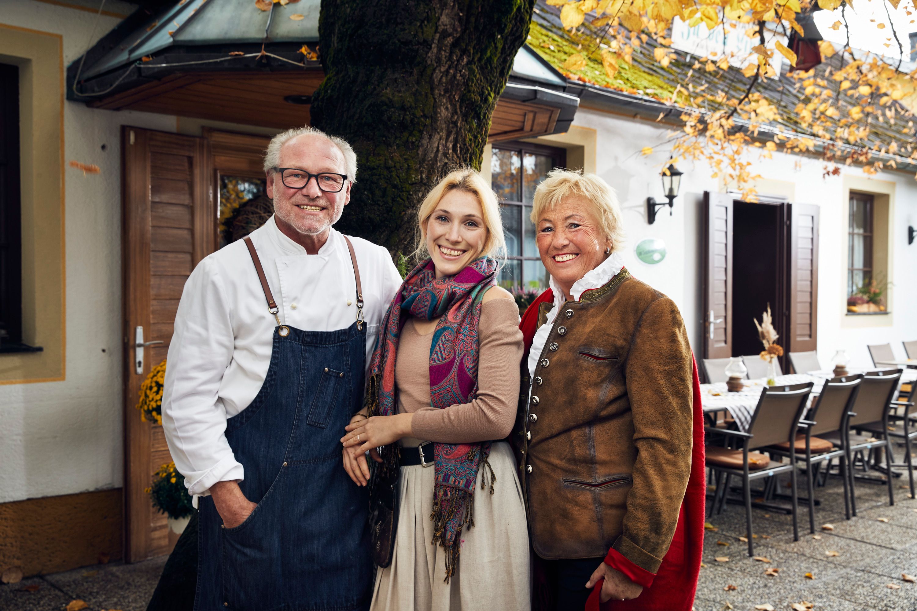 Three people stand smiling in front of a building with autumn leaves.