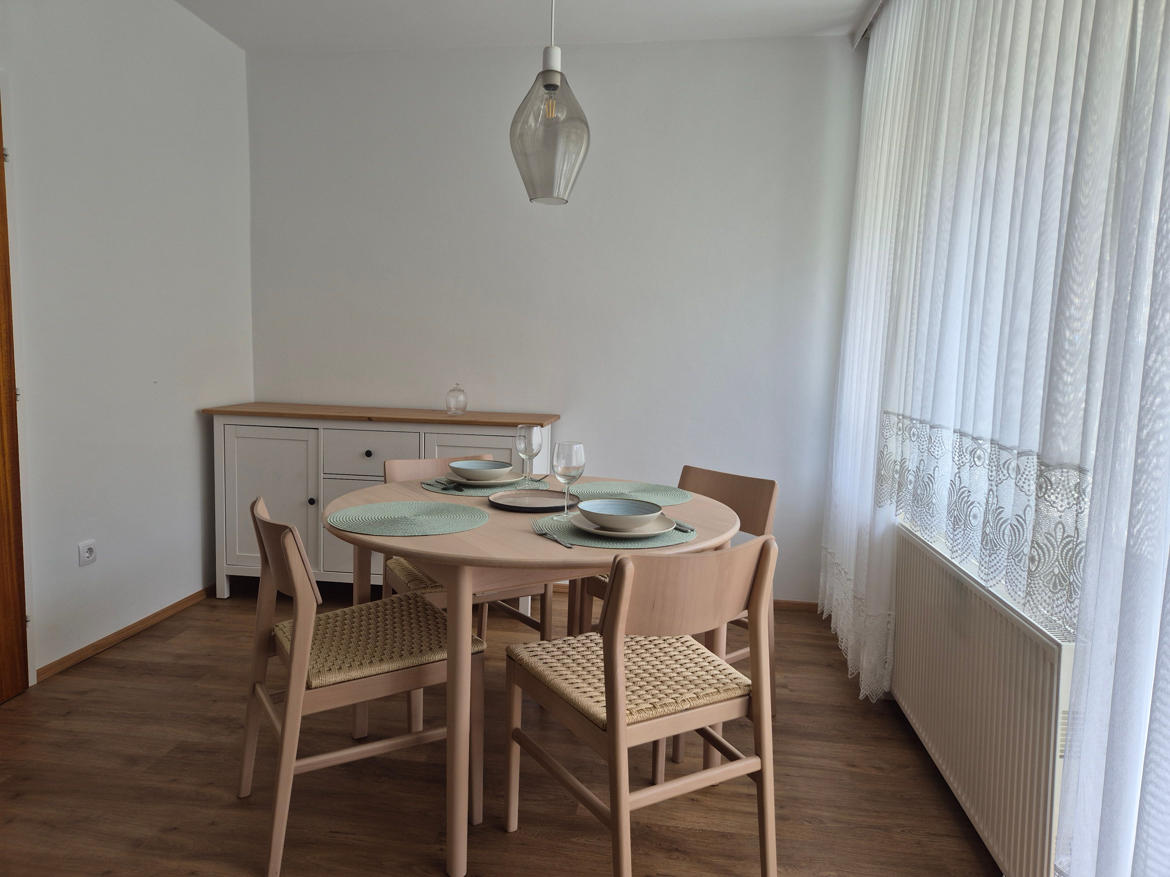 Dining area with round table, four chairs and crockery, next to a window with white curtains.