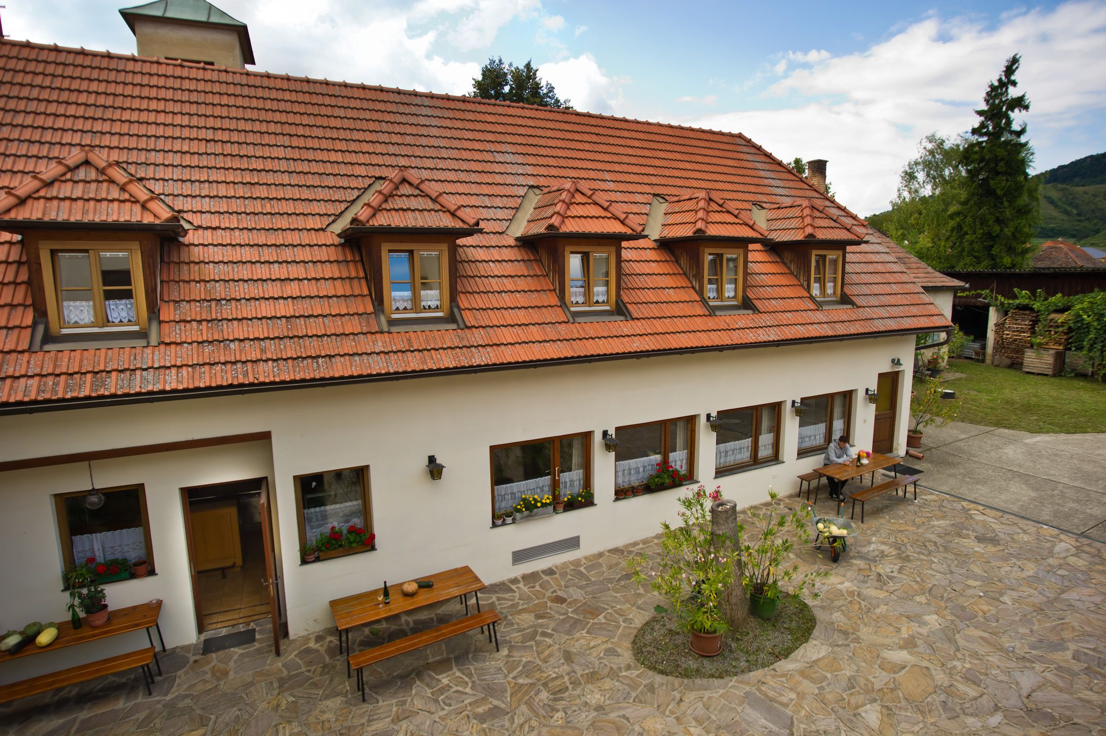 A traditional building with a red tiled roof and skylights, surrounded by a paved courtyard with tables and plants.