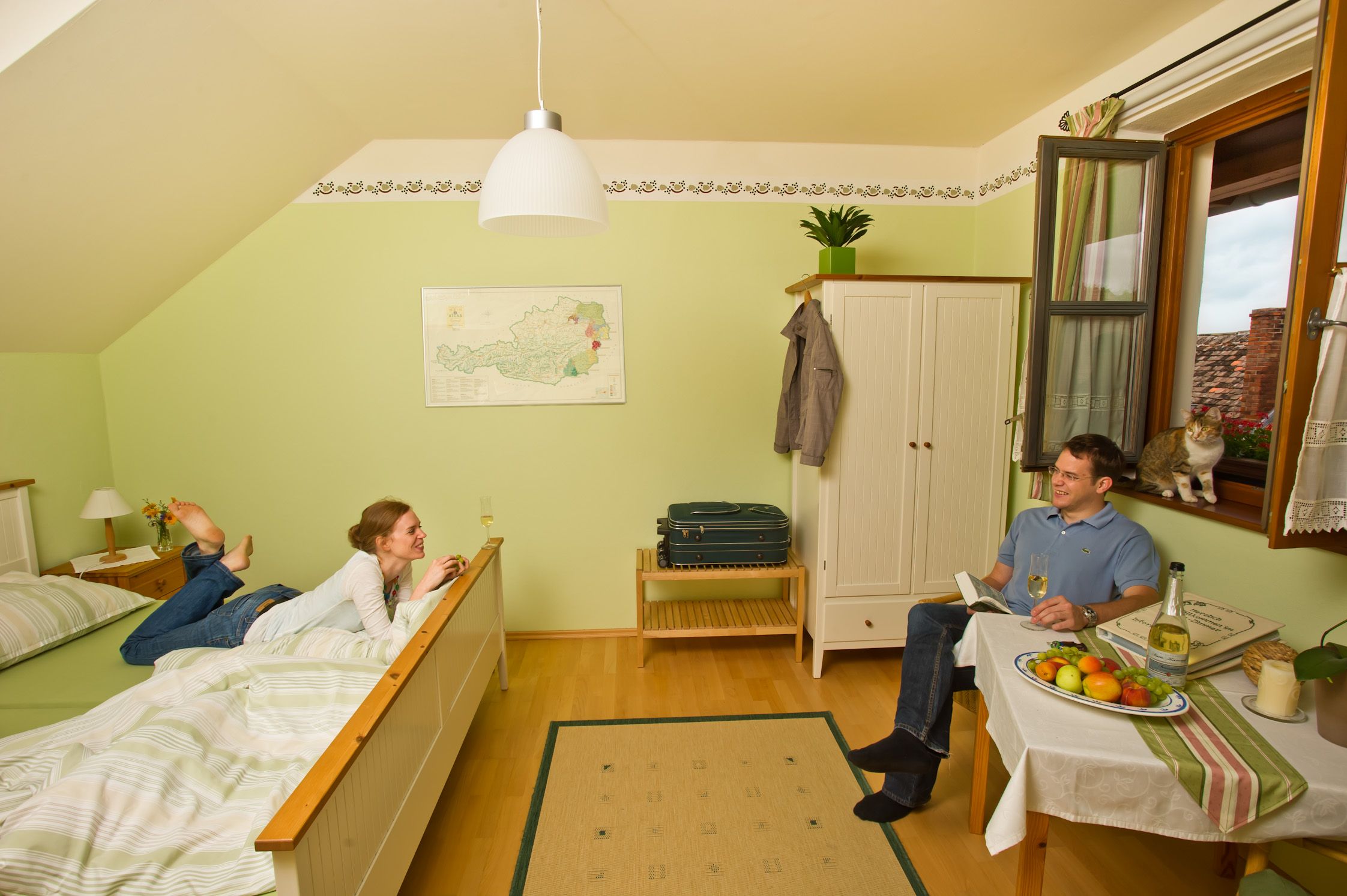A cozy guest room with a couple relaxing. A woman is lying on the bed, a man is sitting at the table. A cat sits by the window.