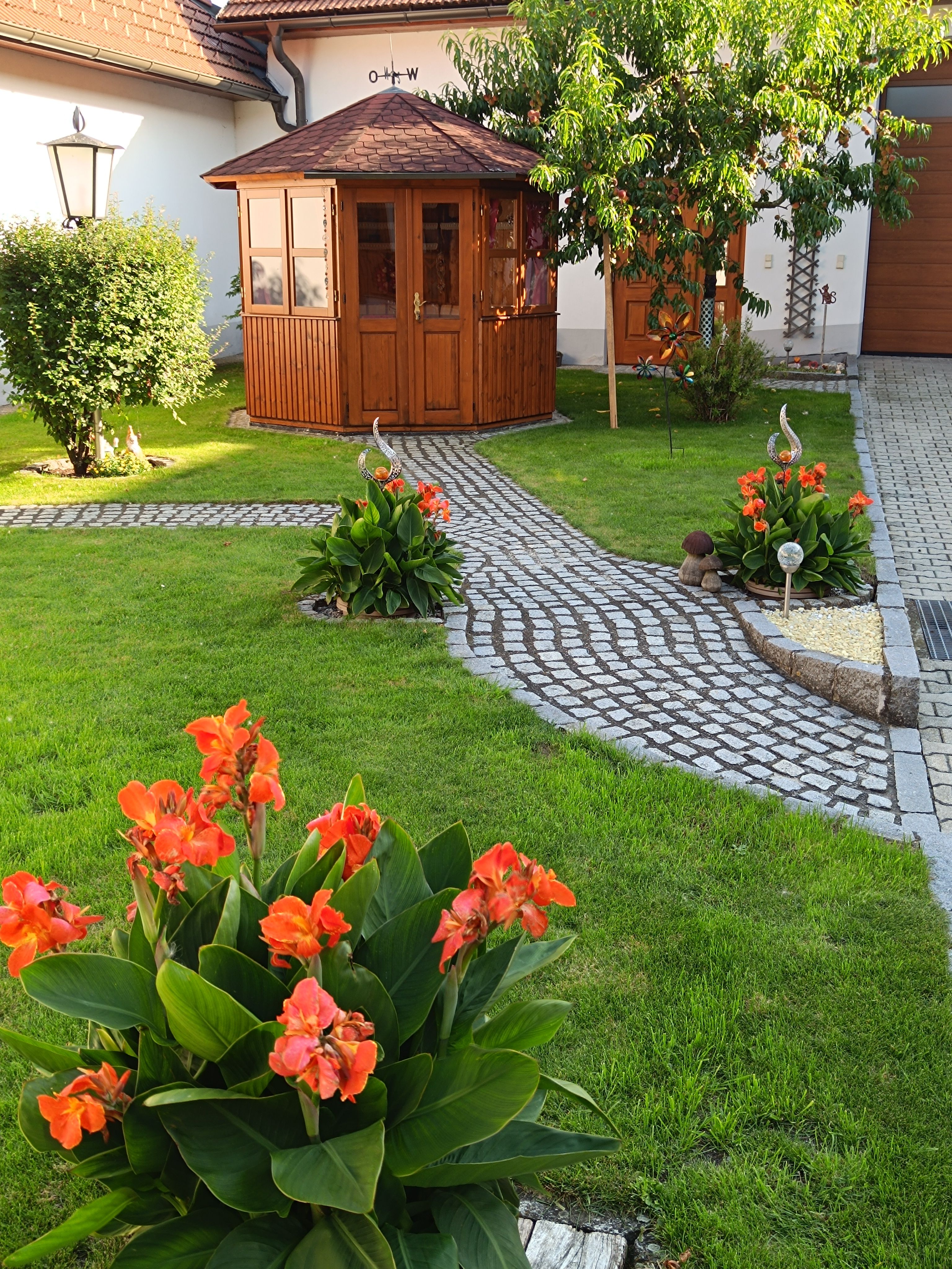 A well-tended garden with a small wooden pavilion, surrounded by green meadows and blooming orange flowers.