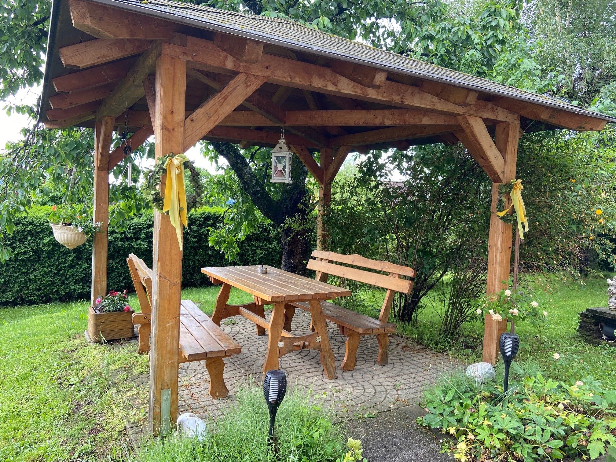 Wooden arbor in the garden with table and benches, surrounded by plants.