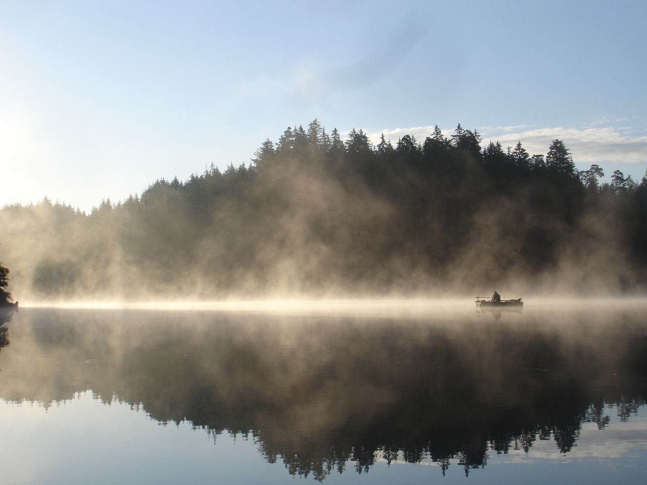 A boat on a misty lake in front of a wooded shore at sunrise.