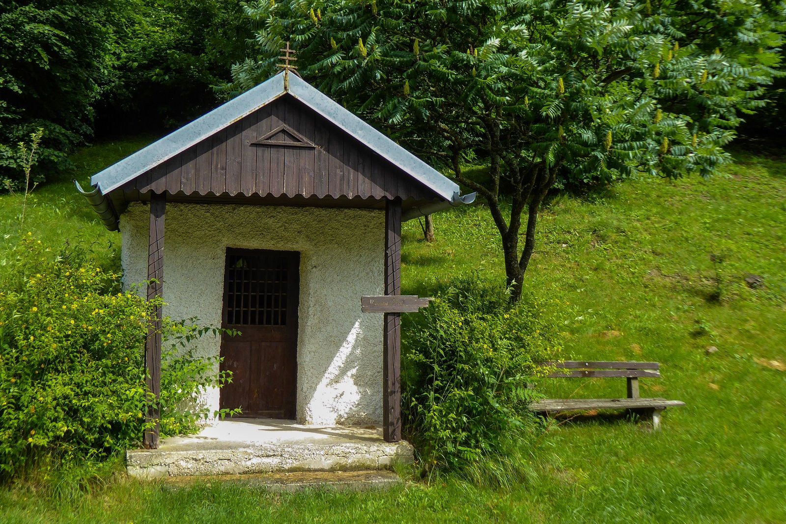 Small chapel with wooden roof and cross, surrounded by green vegetation and a bench.