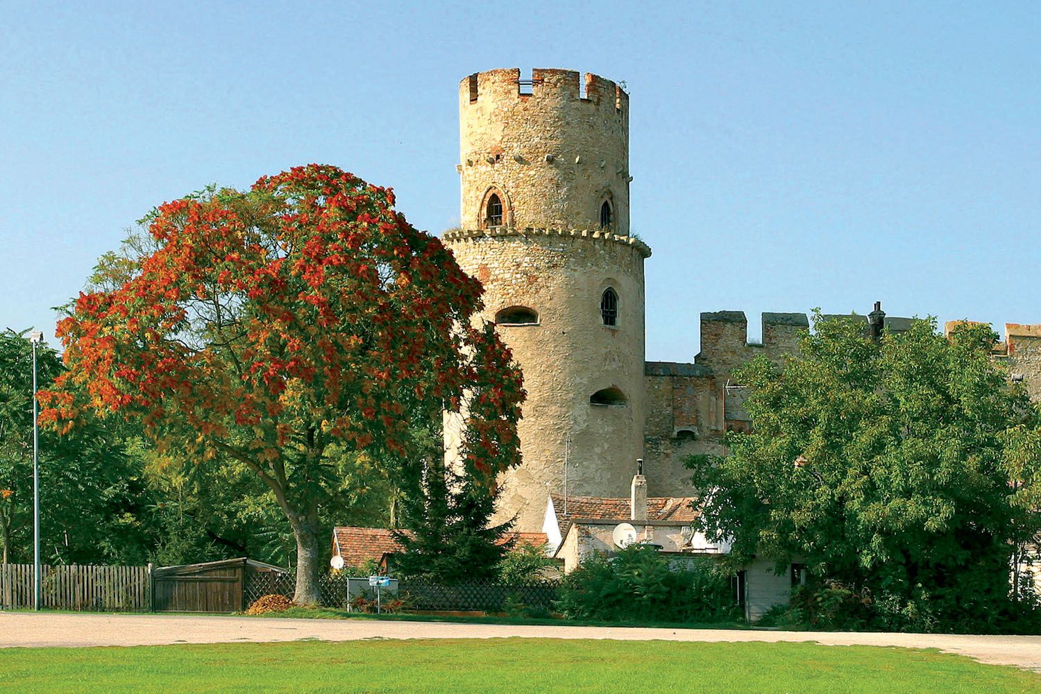 A round stone castle tower next to a tree with red leaves, surrounded by green vegetation and a blue sky.