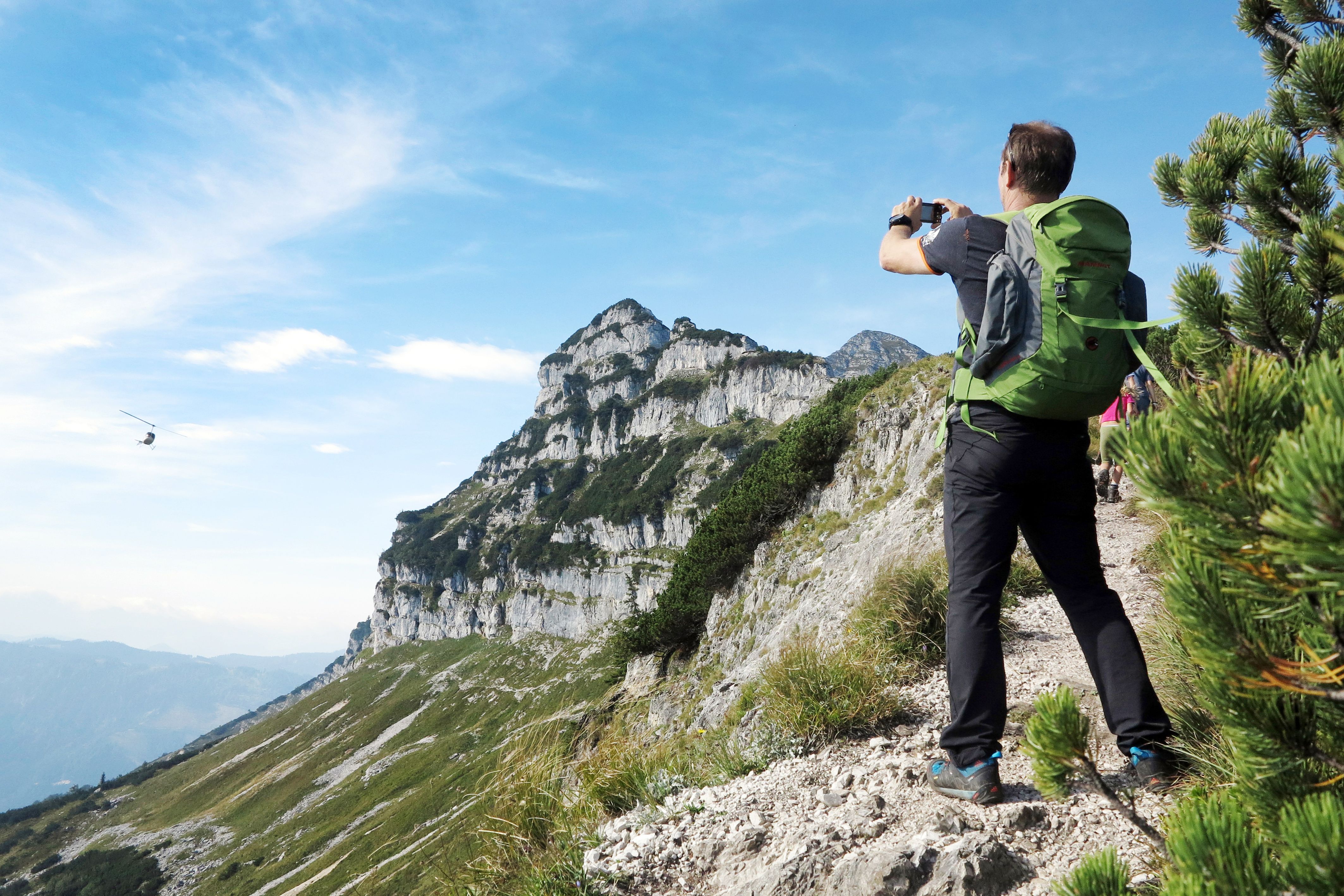 A hiker with a green rucksack photographs a mountain landscape with a helicopter in the sky.